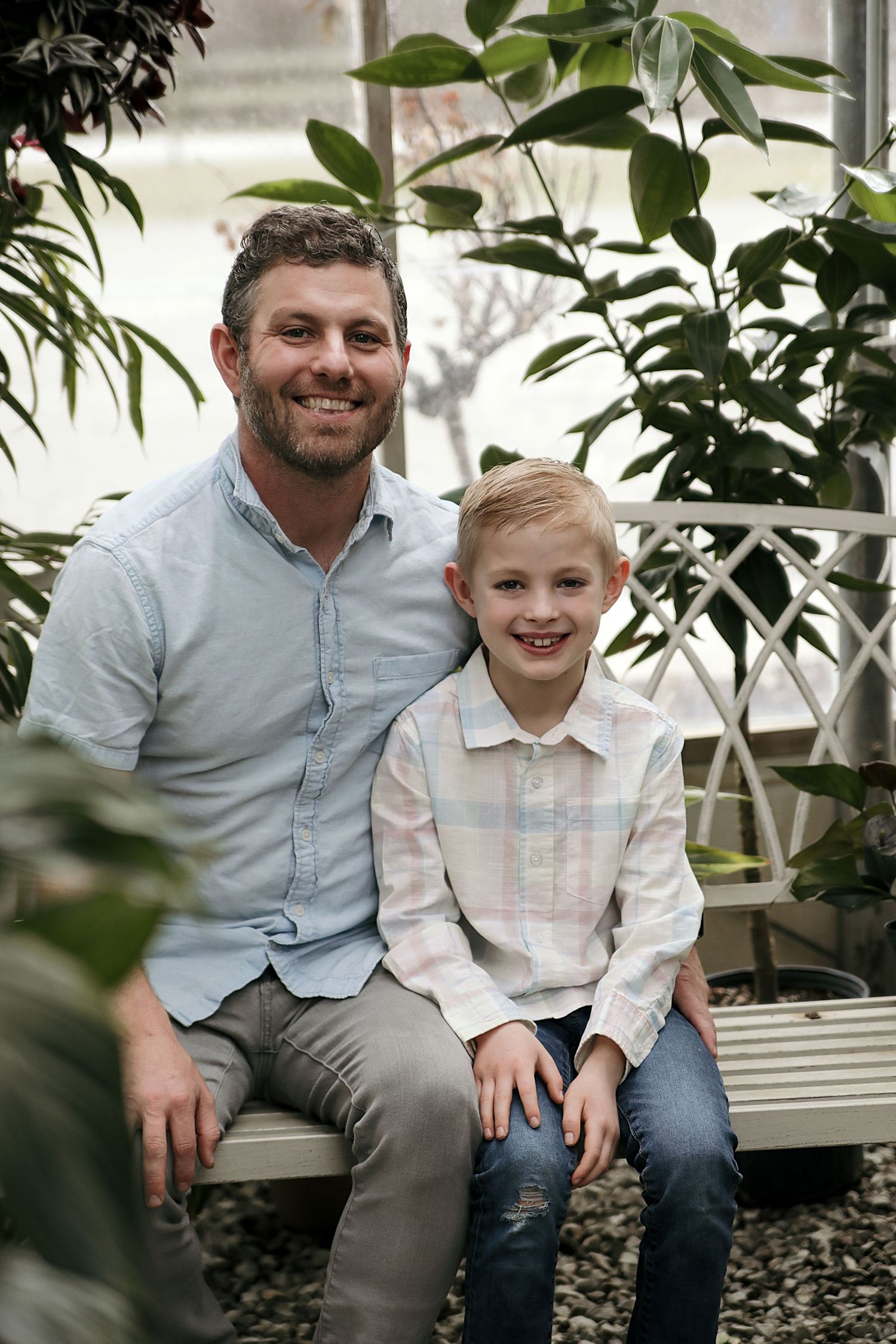 A parent and child sit together on a white bench surrounded by green plants in an indoor garden setting.