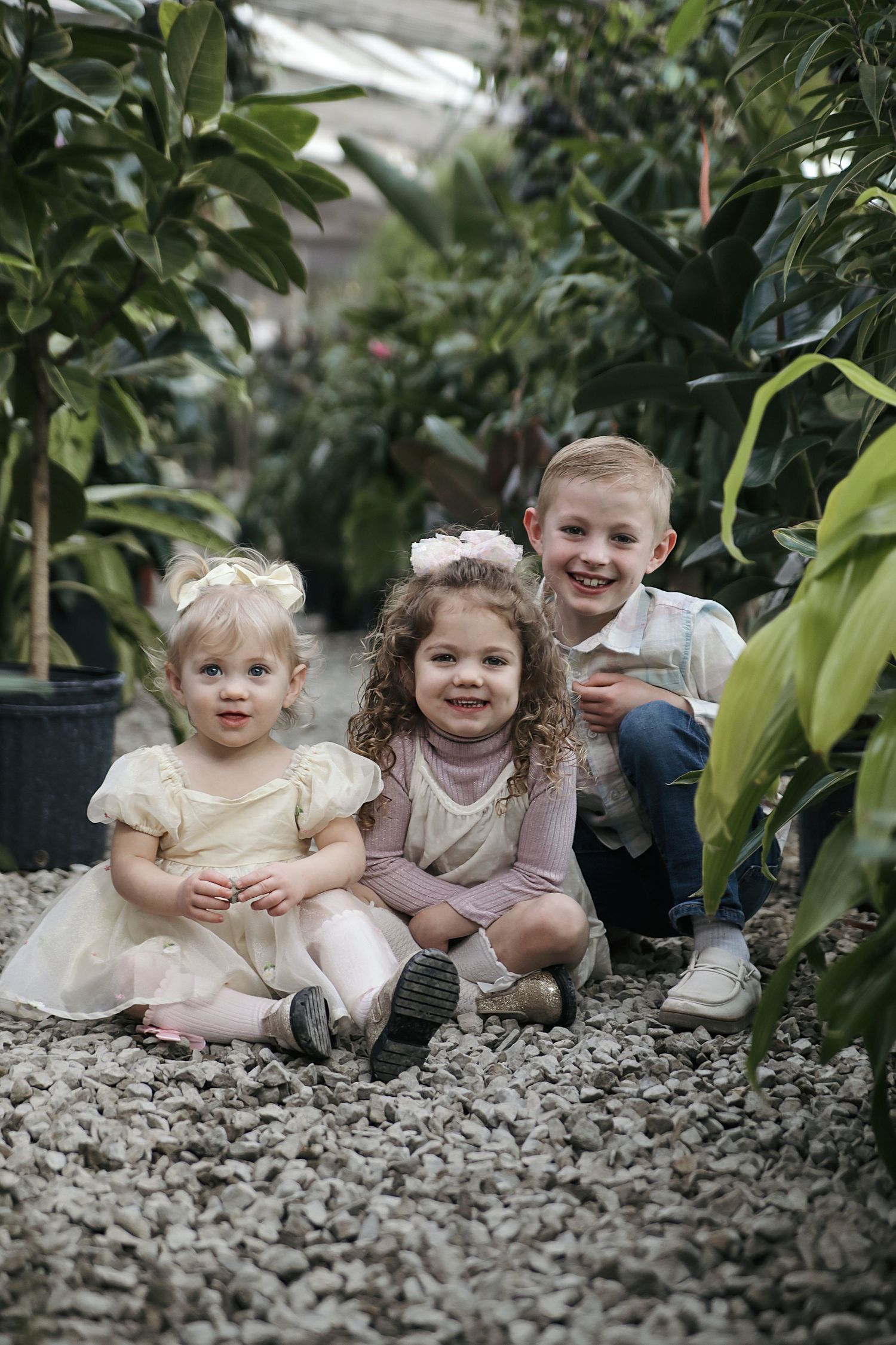 Several children lie on their stomachs on white gravel path surrounded by lush green plants in a greenhouse setting.