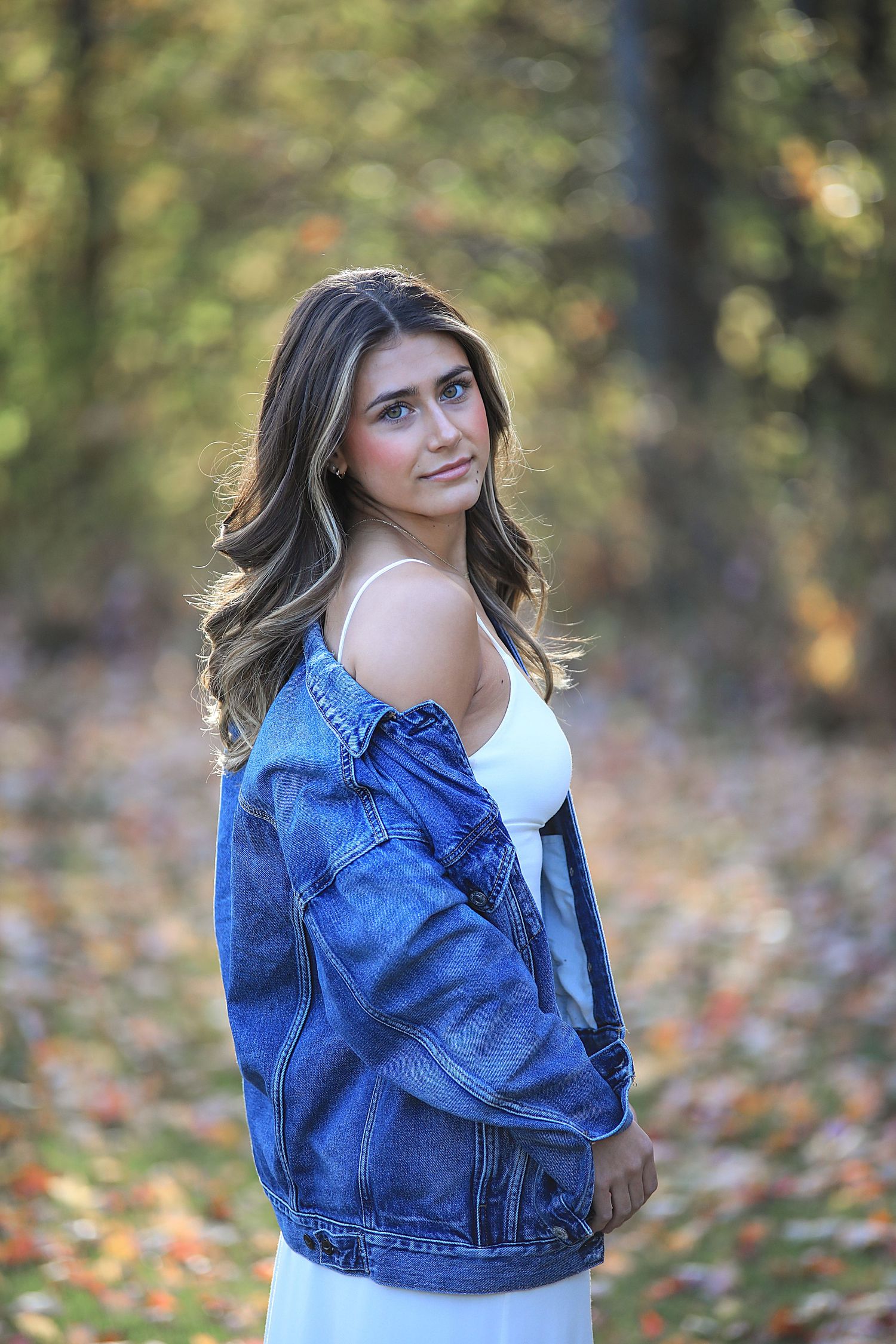 Person wearing an oversized denim jacket and white top poses outdoors with autumn foliage in background.