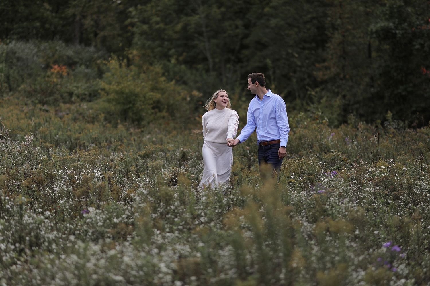 A couple hold hands while walking through a wildflower meadow at dusk.