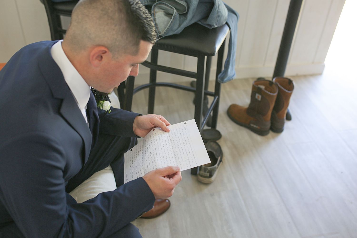 Someone in a navy suit reads from a notebook while sitting in a room with boots visible in the background.