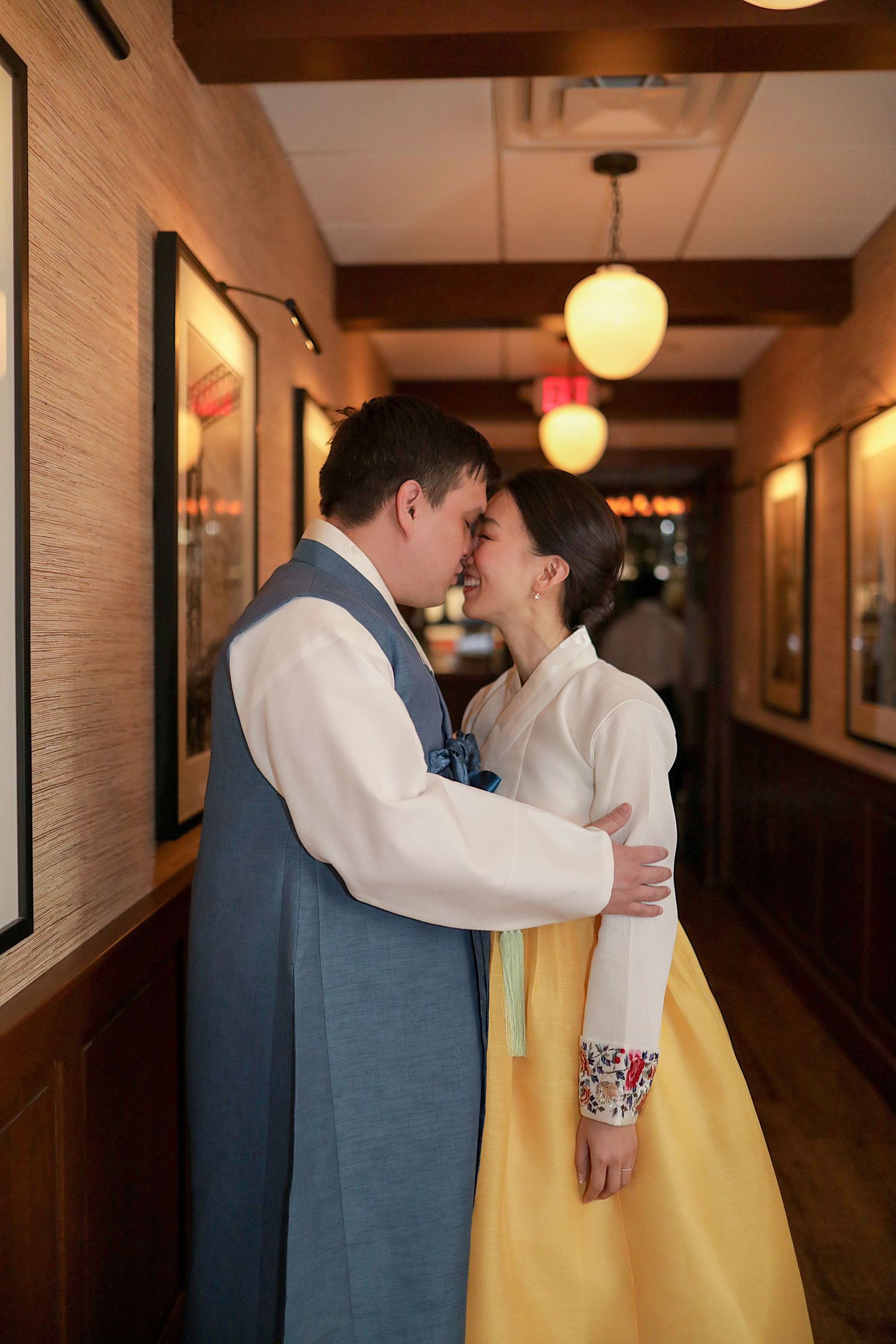 A couple in traditional Korean hanbok embraces in softly lit hallway with yellow paper lanterns overhead.