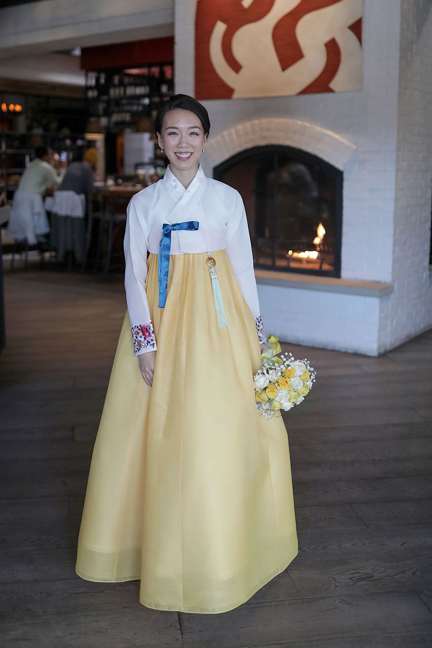 Someone wearing a traditional Korean hanbok with a white top and yellow skirt holds a floral bouquet in an indoor setting.
