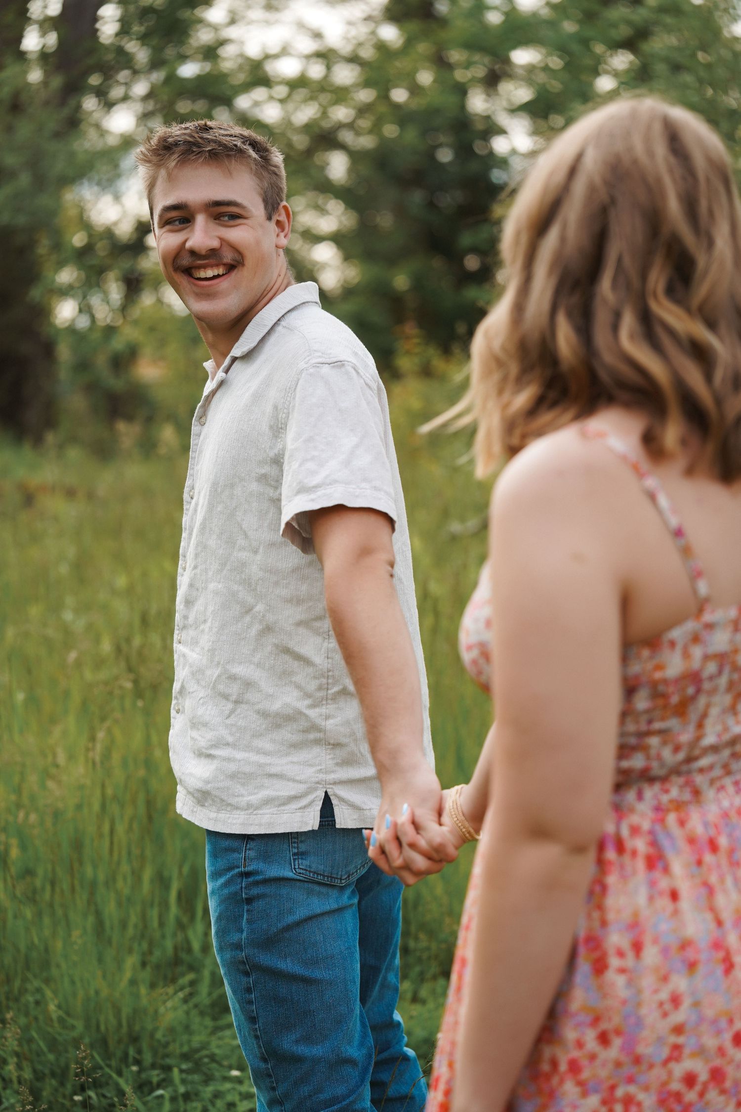 A couple holds hands while walking through tall grass on a sunny summer day, laughing and enjoying each other's company.