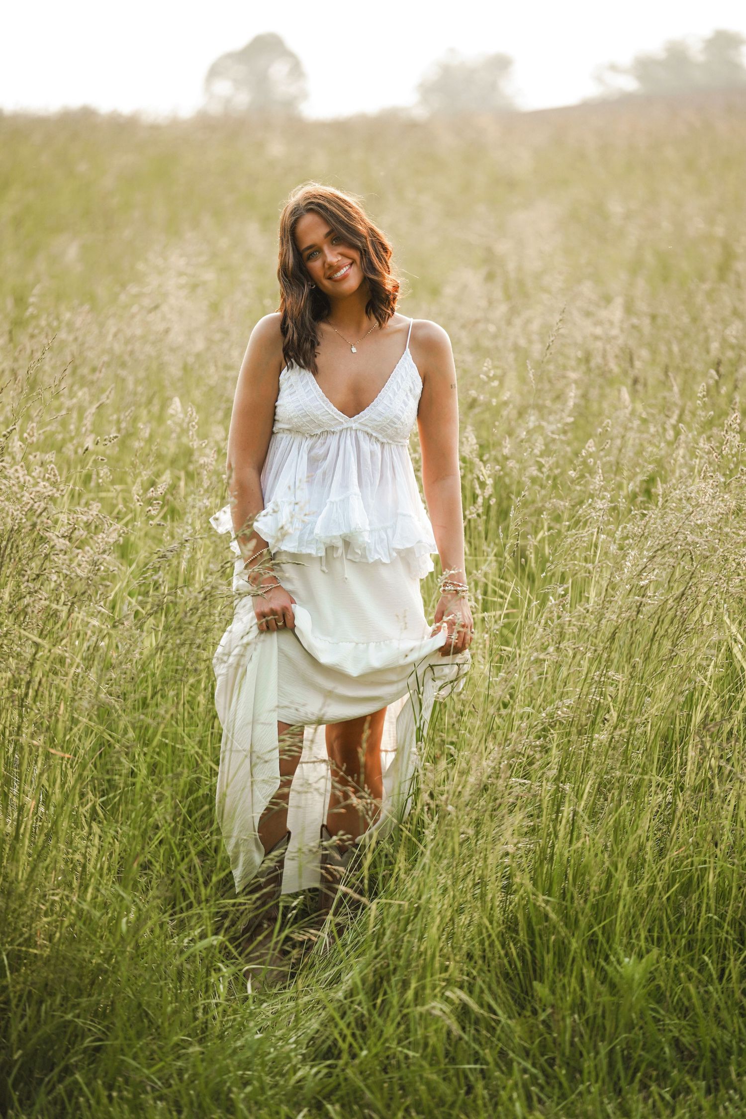 A figure in a flowing white summer dress stands in a sunlit meadow of tall grass during golden hour.