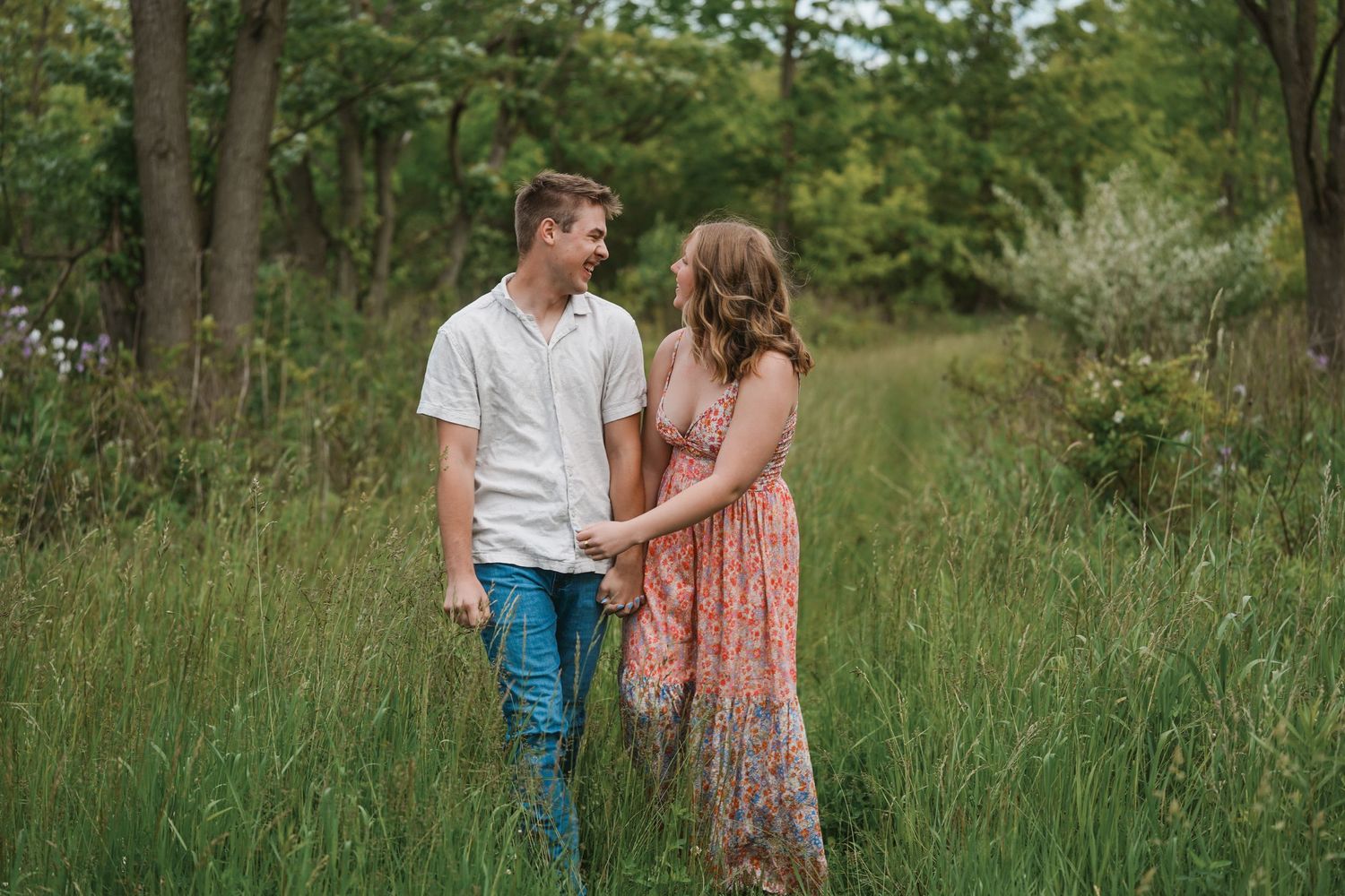 A couple in casual attire hold hands while standing in a green meadow surrounded by trees on a summer day.