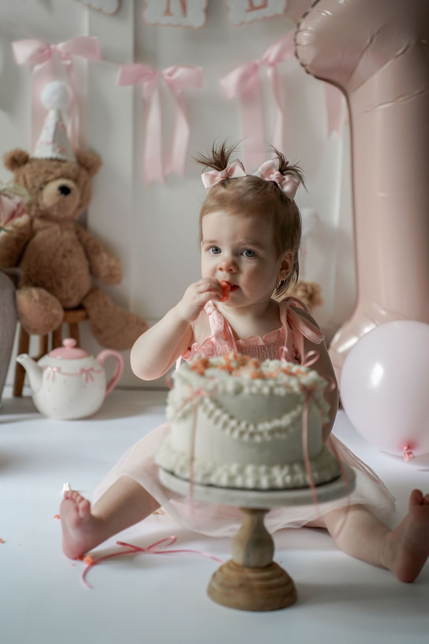 A first birthday party setup with a decorated cake, pink ribbons, teddy bear, and baby in a pink dress sitting on white floor.