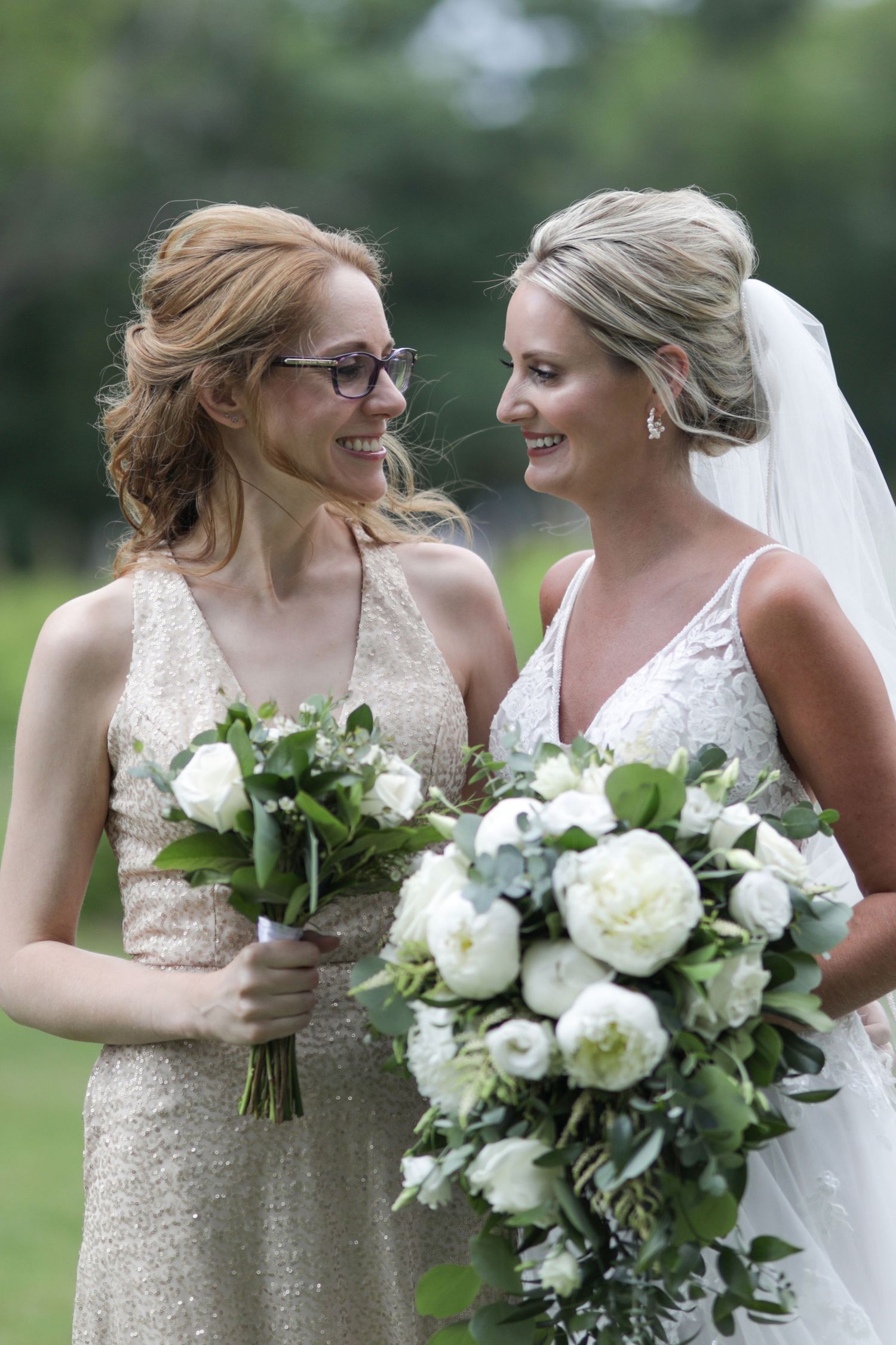 Wedding party members share joyful smiles while holding elegant white floral bouquets in an outdoor setting.