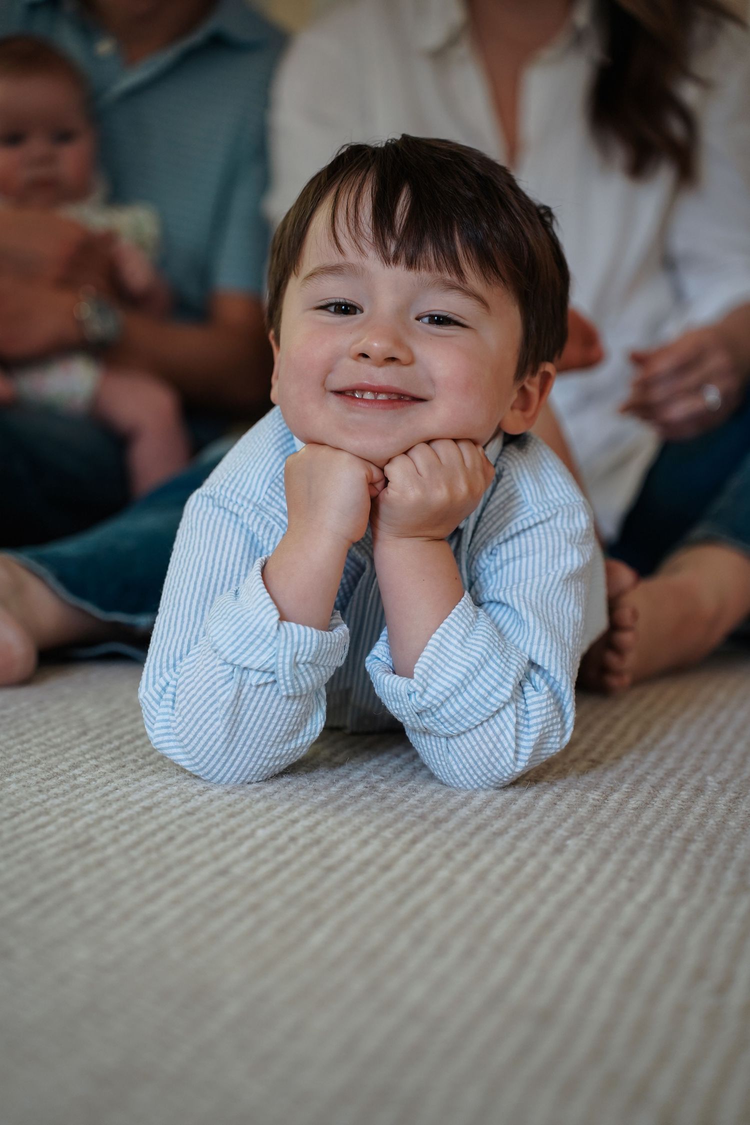 Child lying on gray carpet wearing light blue button-up shirt rests chin on hands with playful expressions.