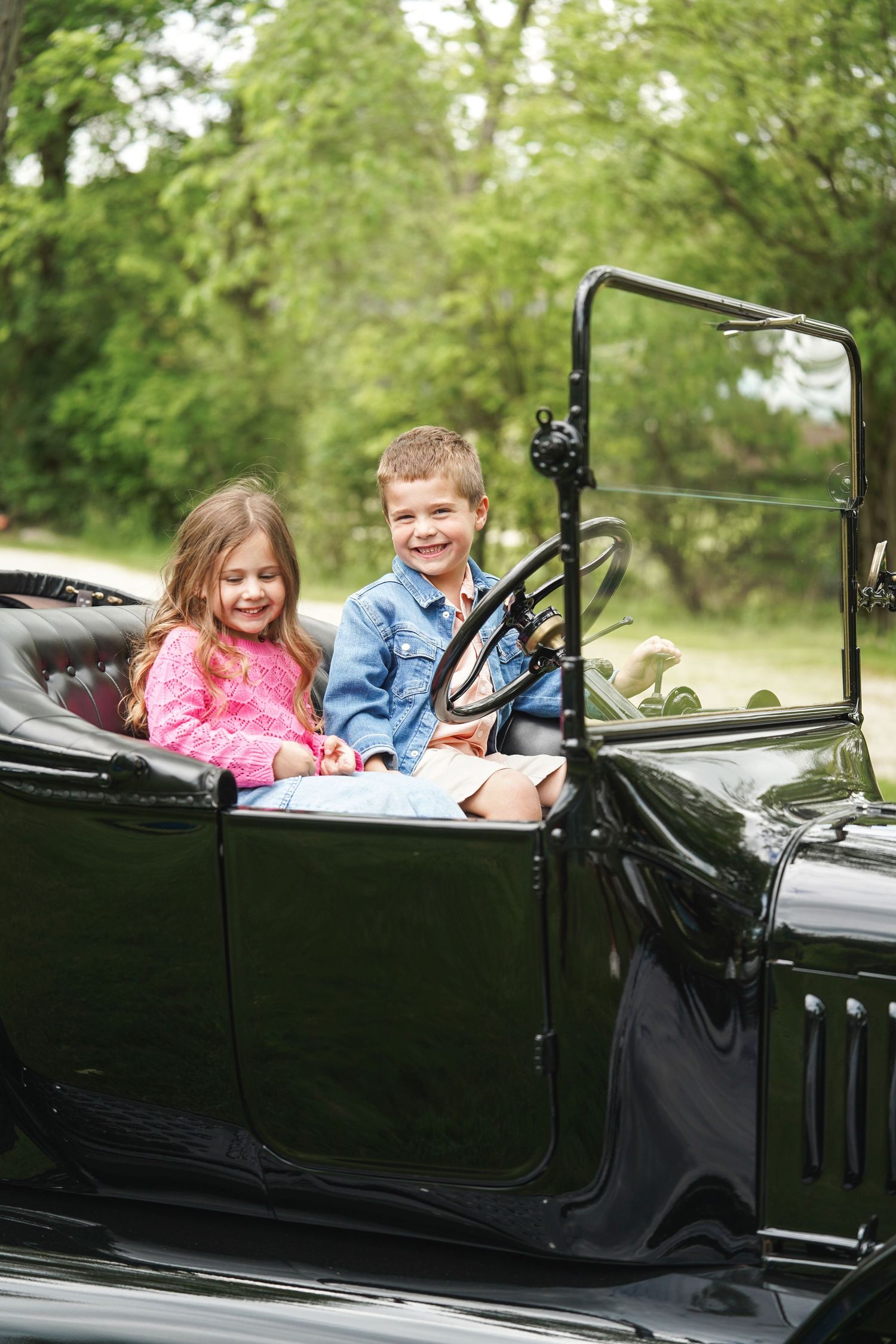 Two children sitting in a vintage black convertible car smiling and enjoying a ride outdoors.