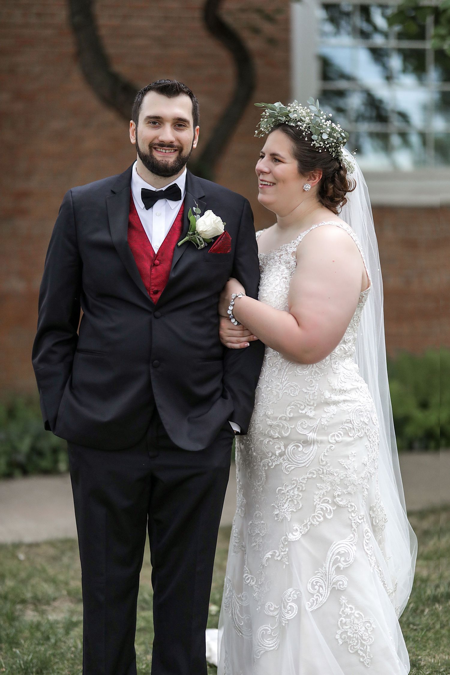 A newlywed couple stands together outdoors, wearing a dark tuxedo with red vest and white lace wedding gown.