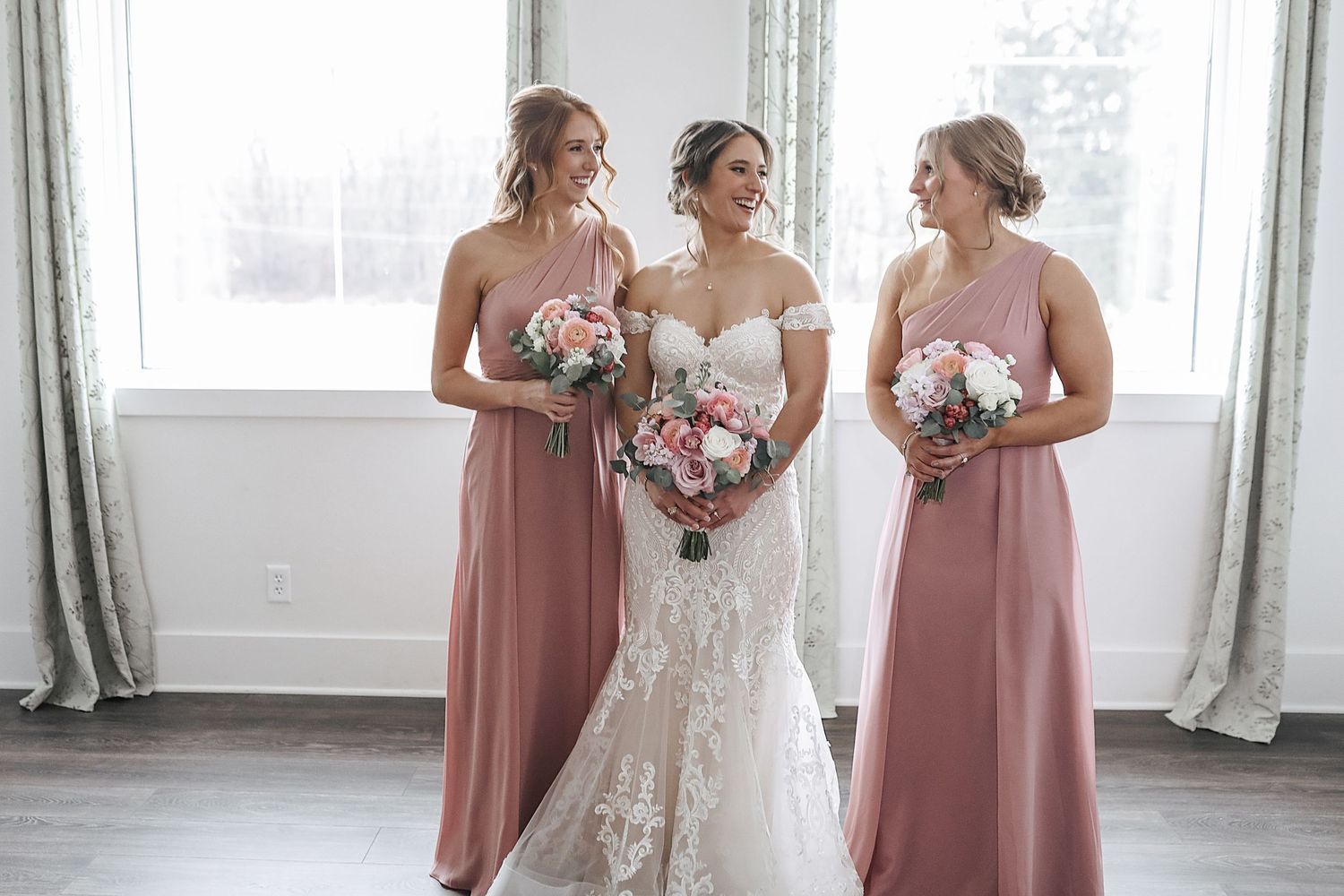 Wedding party in dusty rose bridesmaid dresses and white lace bridal gown pose together in bright indoor venue.