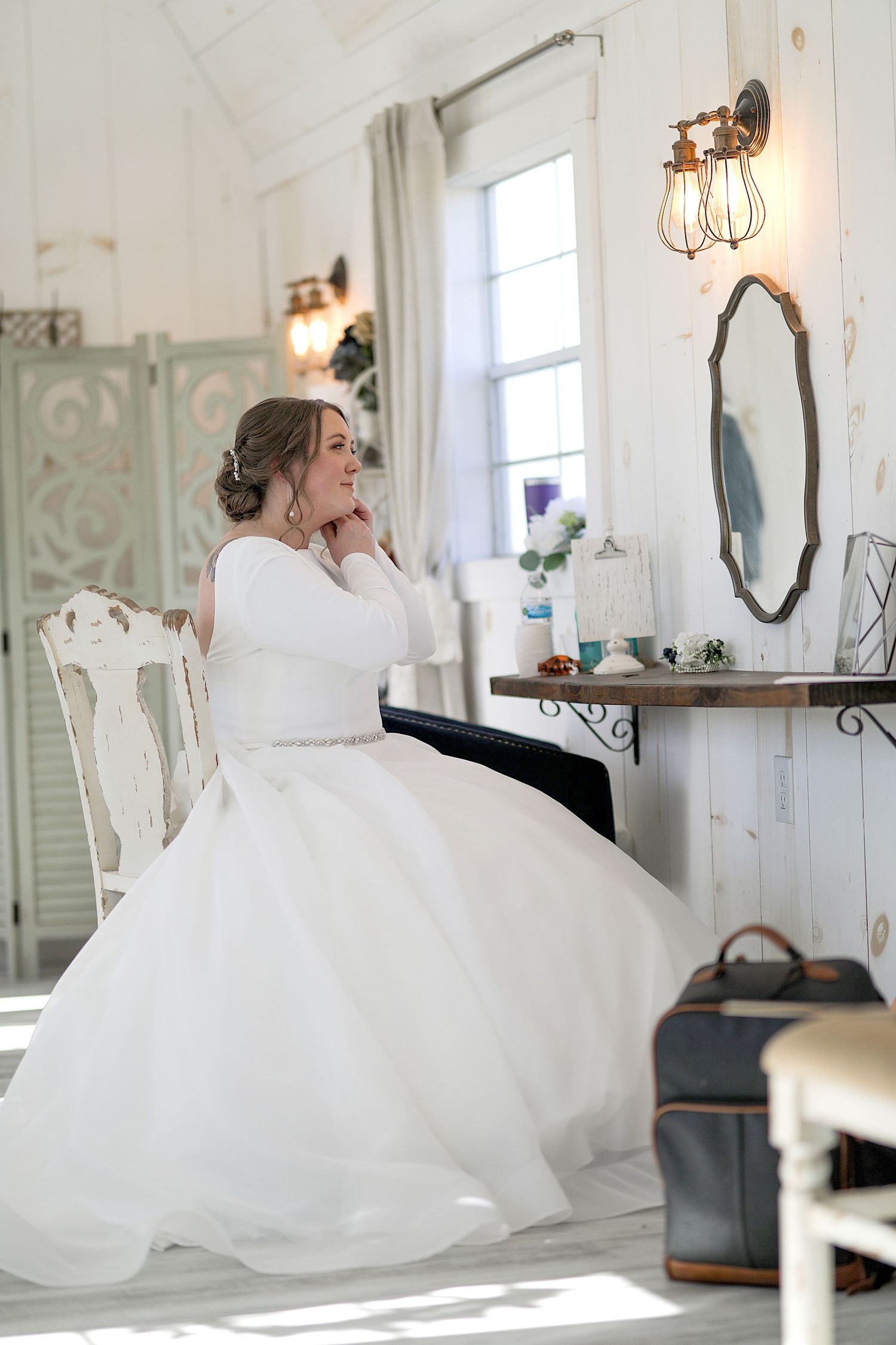 Bride in white gown relaxing on vintage furniture in a light-filled bridal suite.