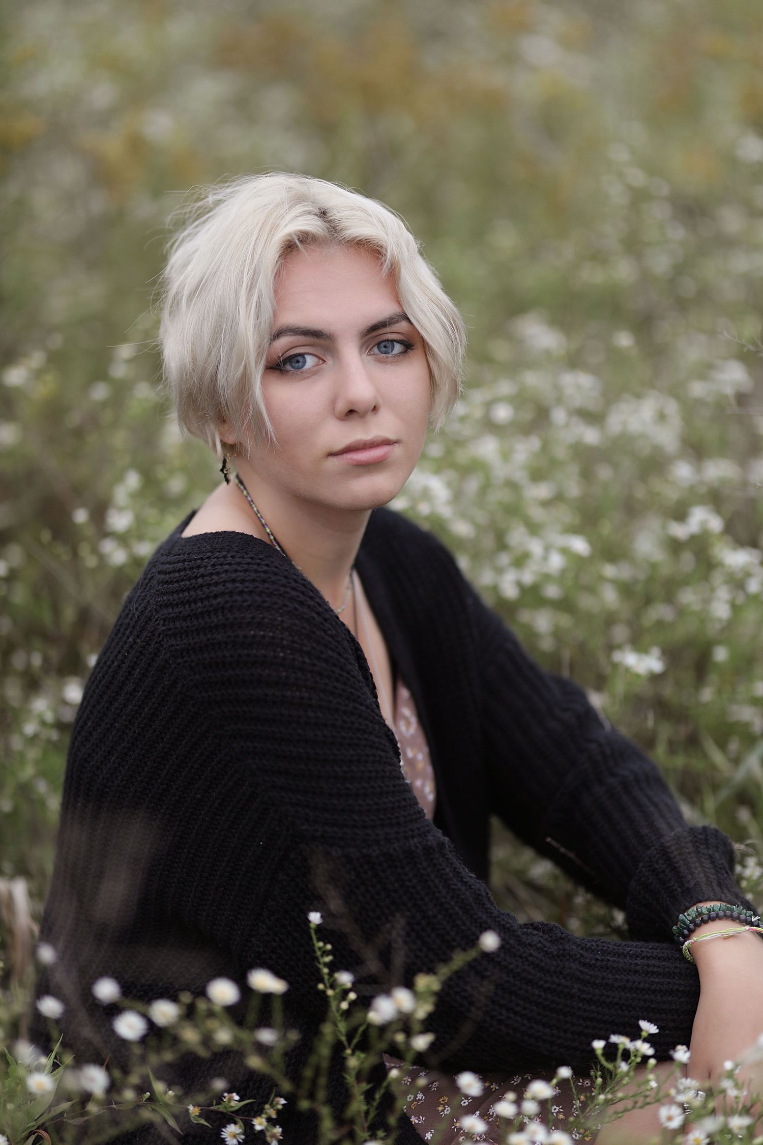 A portrait in a meadow of white wildflowers features a person with platinum blonde hair wearing a black knit sweater.