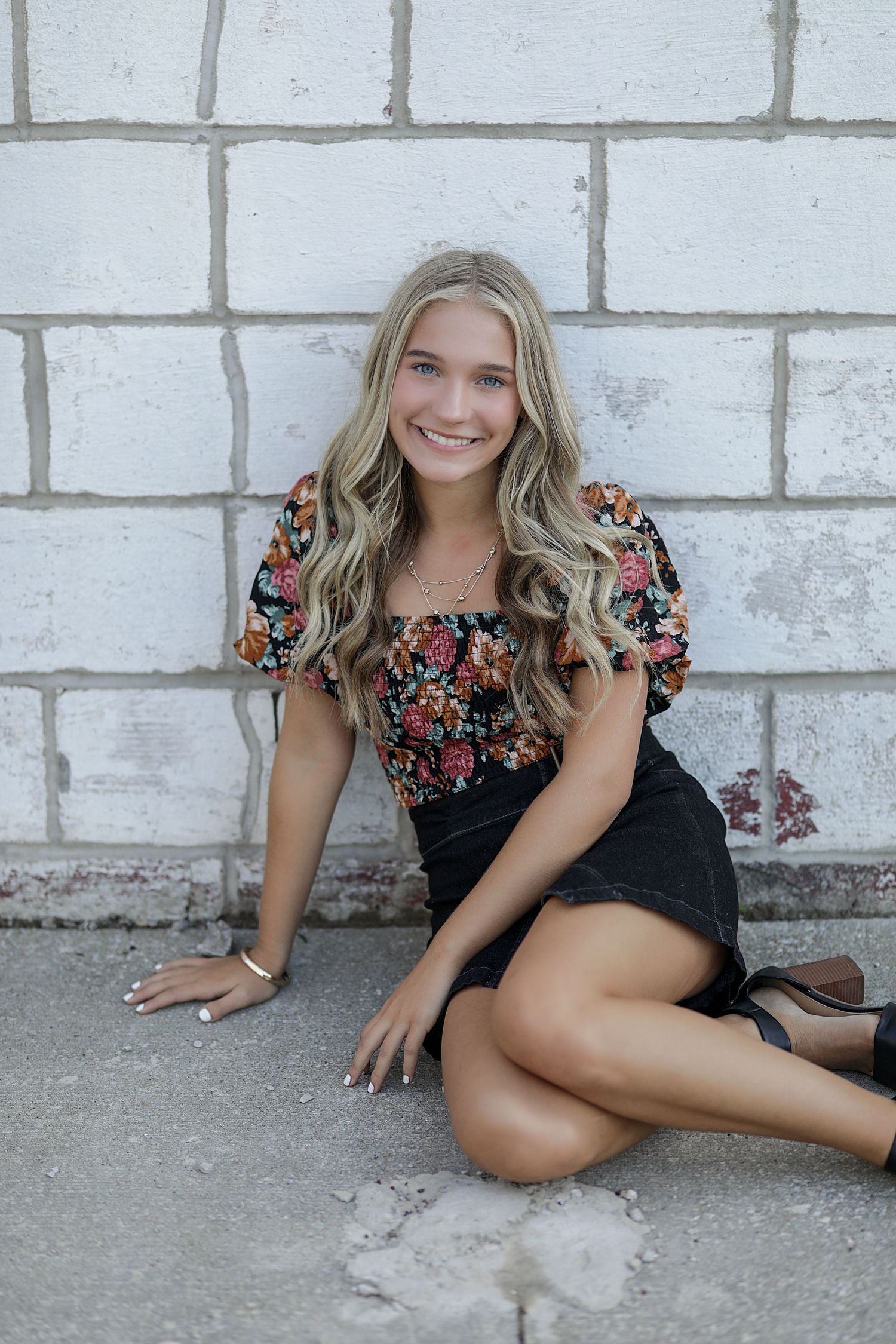 A person in a black floral dress poses casually against a white brick wall in a sequence of four similar photos.