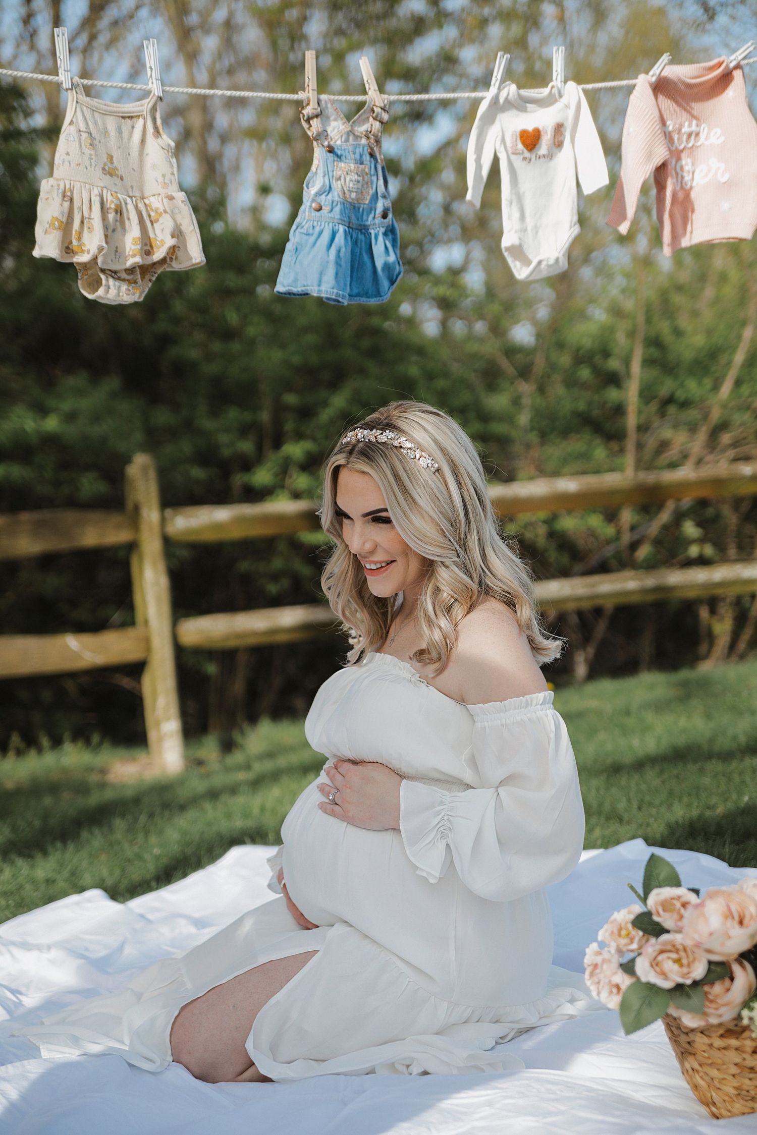 Maternity photo session with vintage style baby clothes on clothesline and soft blanket in outdoor garden setting.