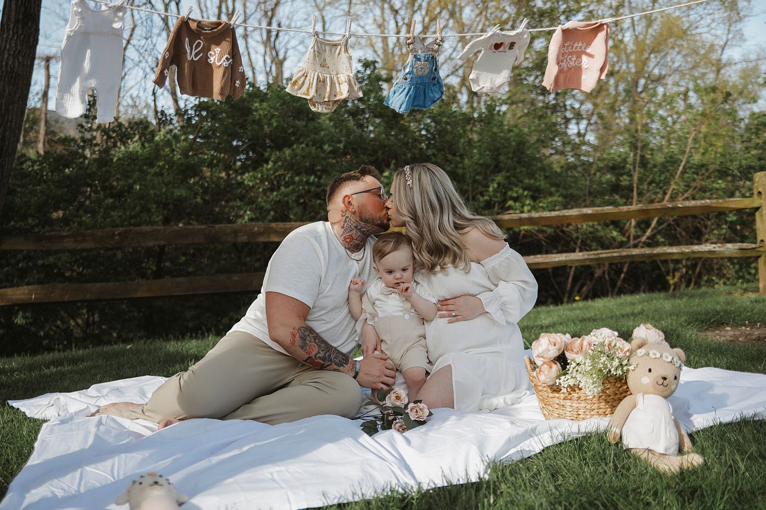 A couple sits on a white blanket at an outdoor maternity photoshoot with teddy bears and hanging decorations above.