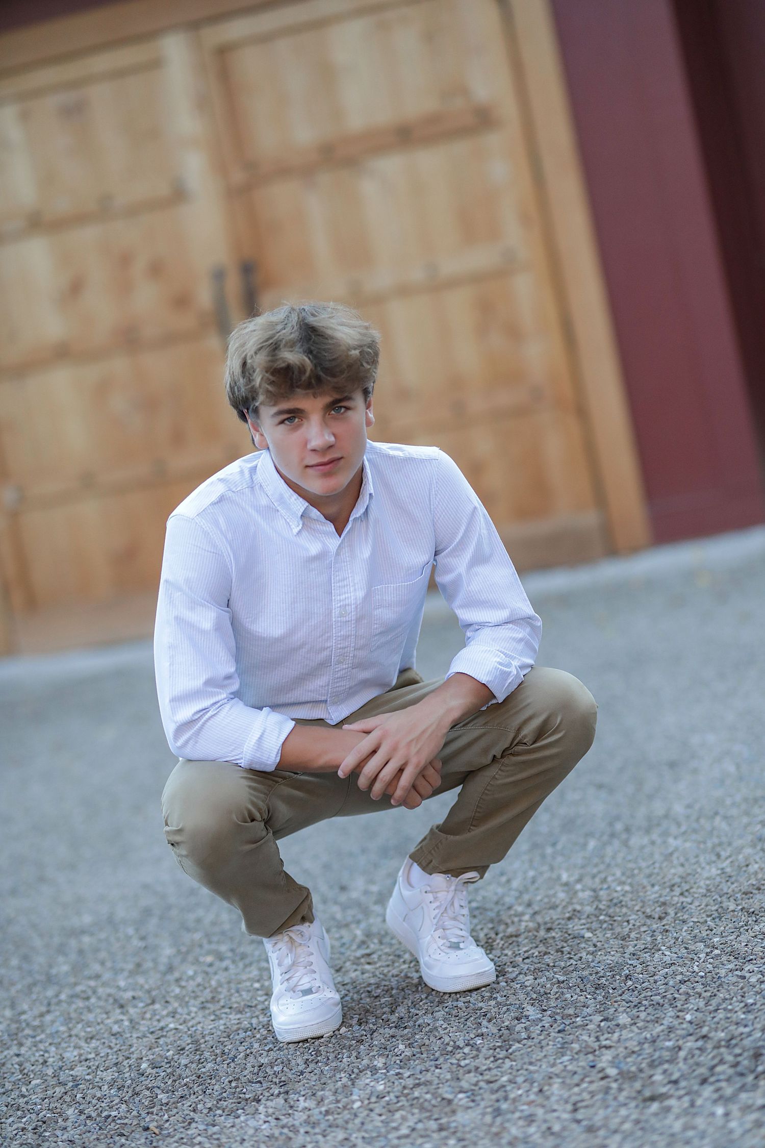 Young person in white button-up shirt and khaki pants crouches on gravel surface near wooden doors.