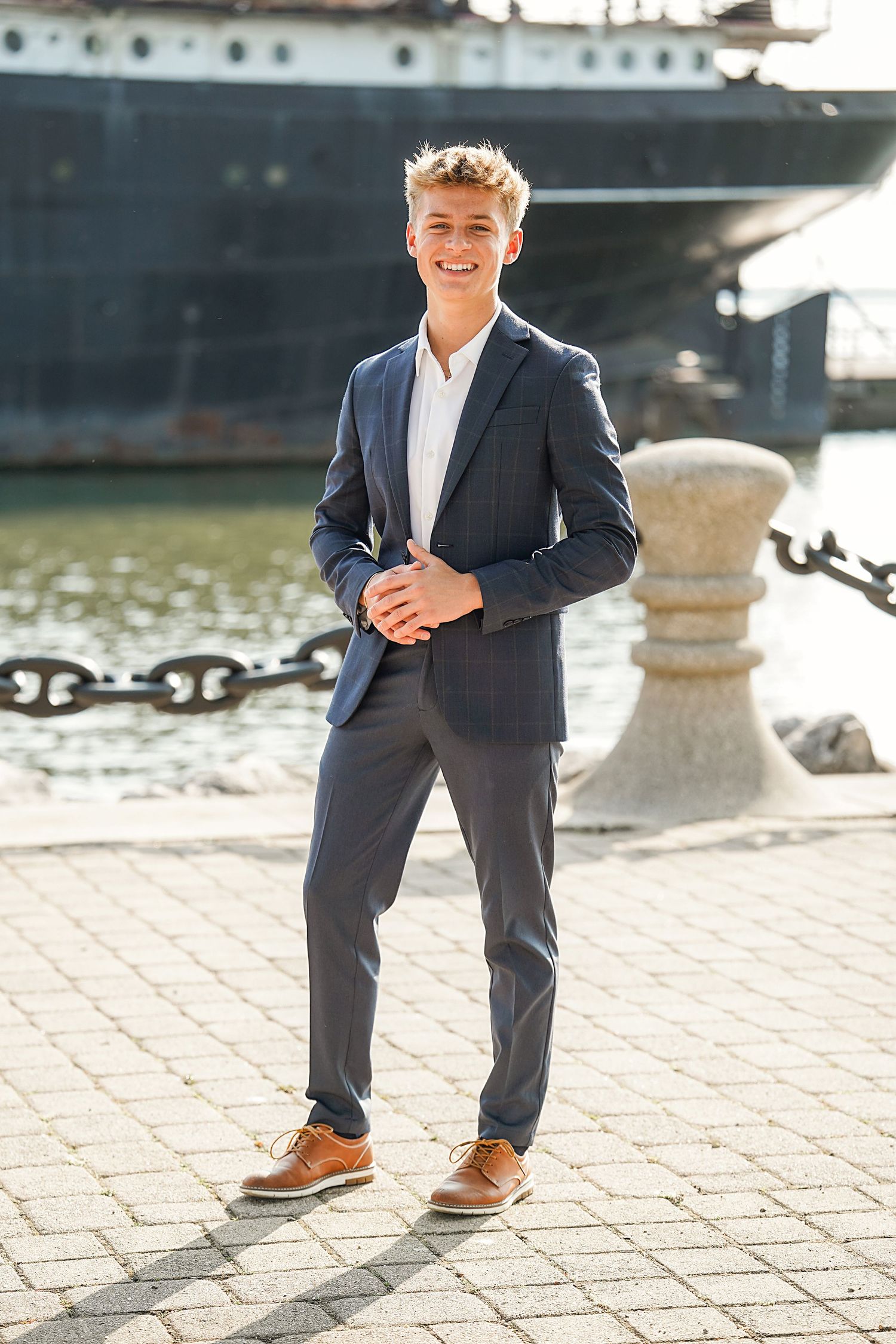 Well-dressed person in a navy suit and brown shoes stands on a waterfront promenade near a large ship.