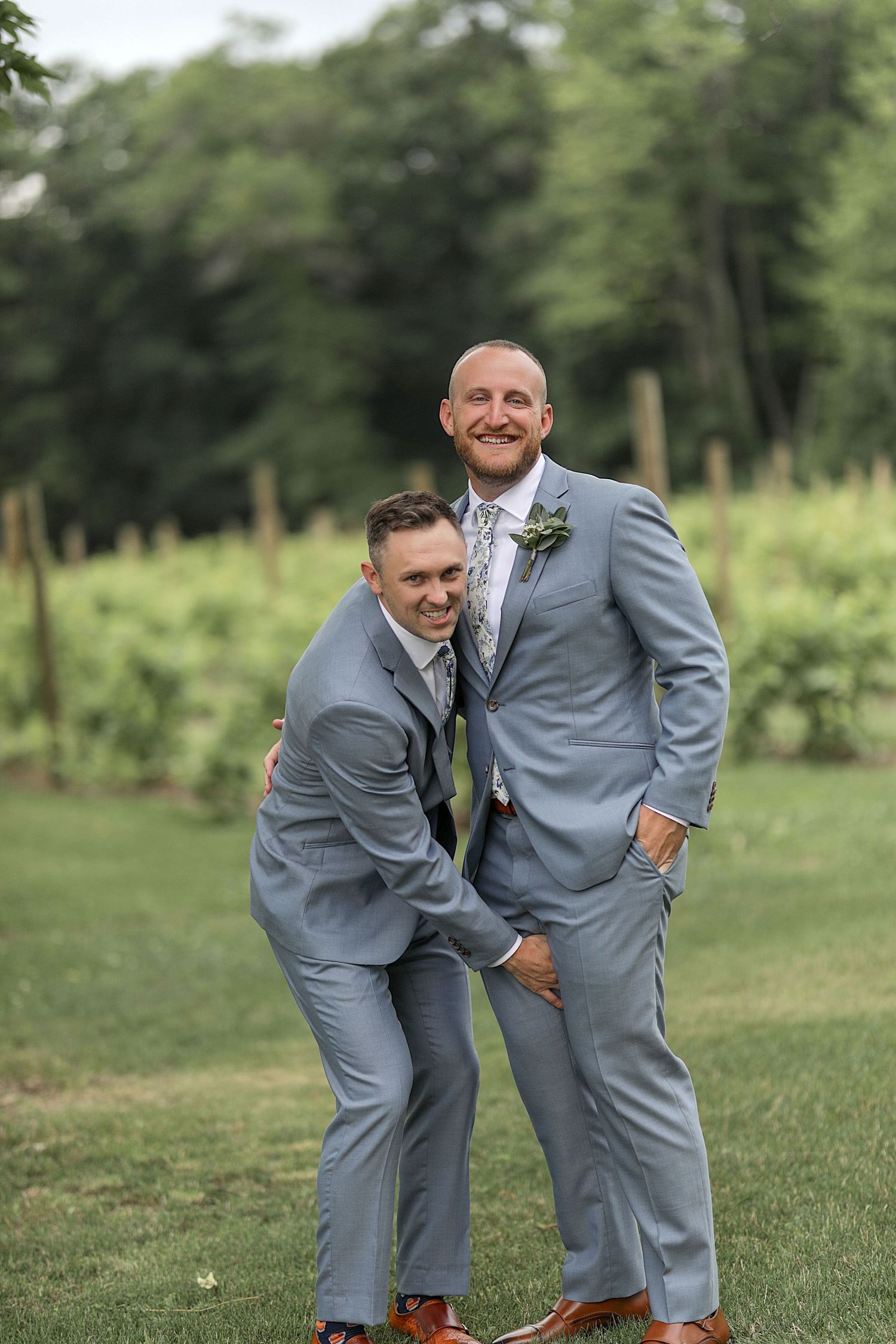Two people in matching gray suits share a playful moment during wedding photos.