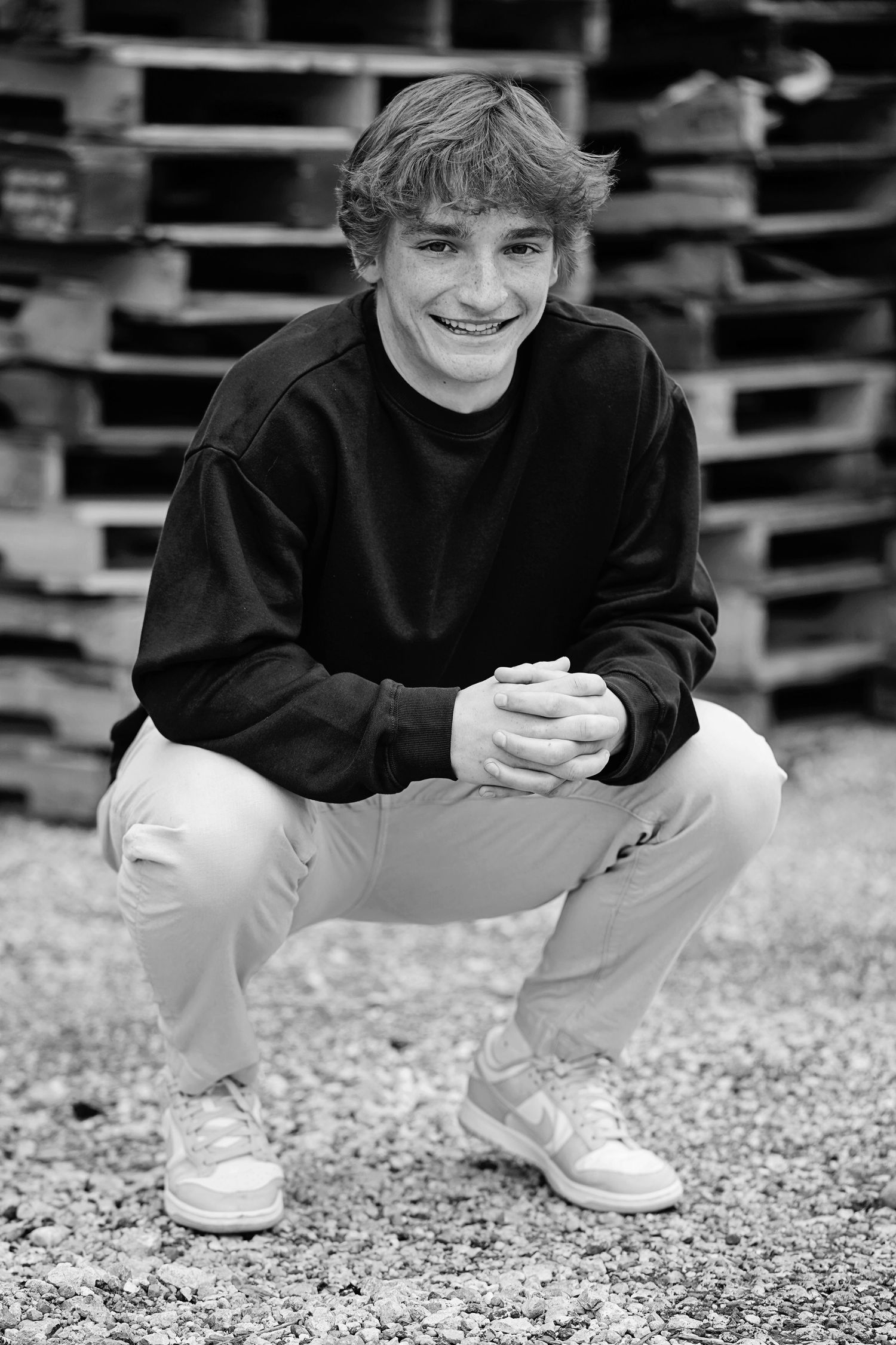 Black and white portrait of someone in casual attire crouching outdoors wearing a dark sweater and light-colored pants.