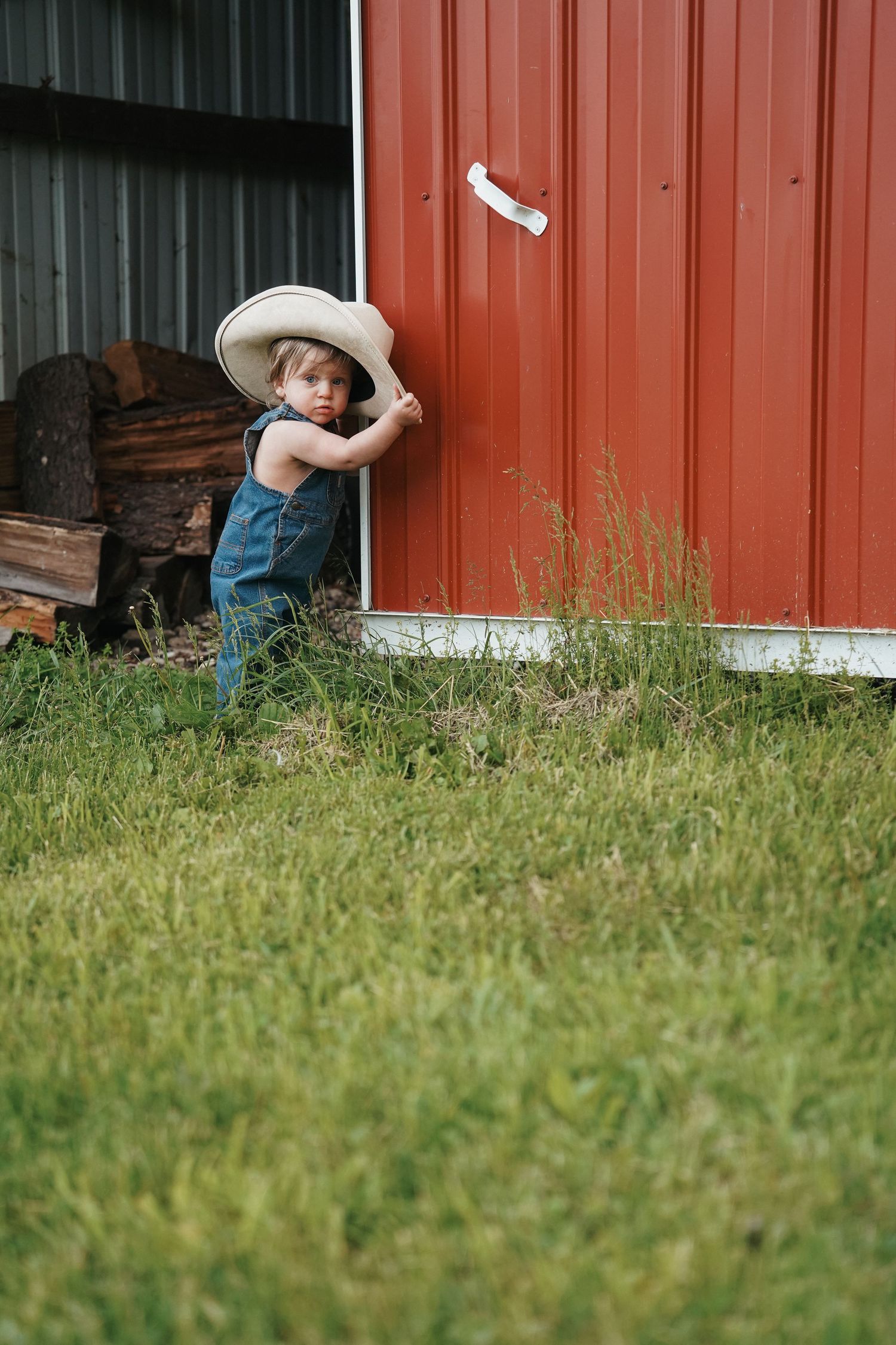 Child in overalls plays hide and seek around a red barn wall in a rural farm setting.