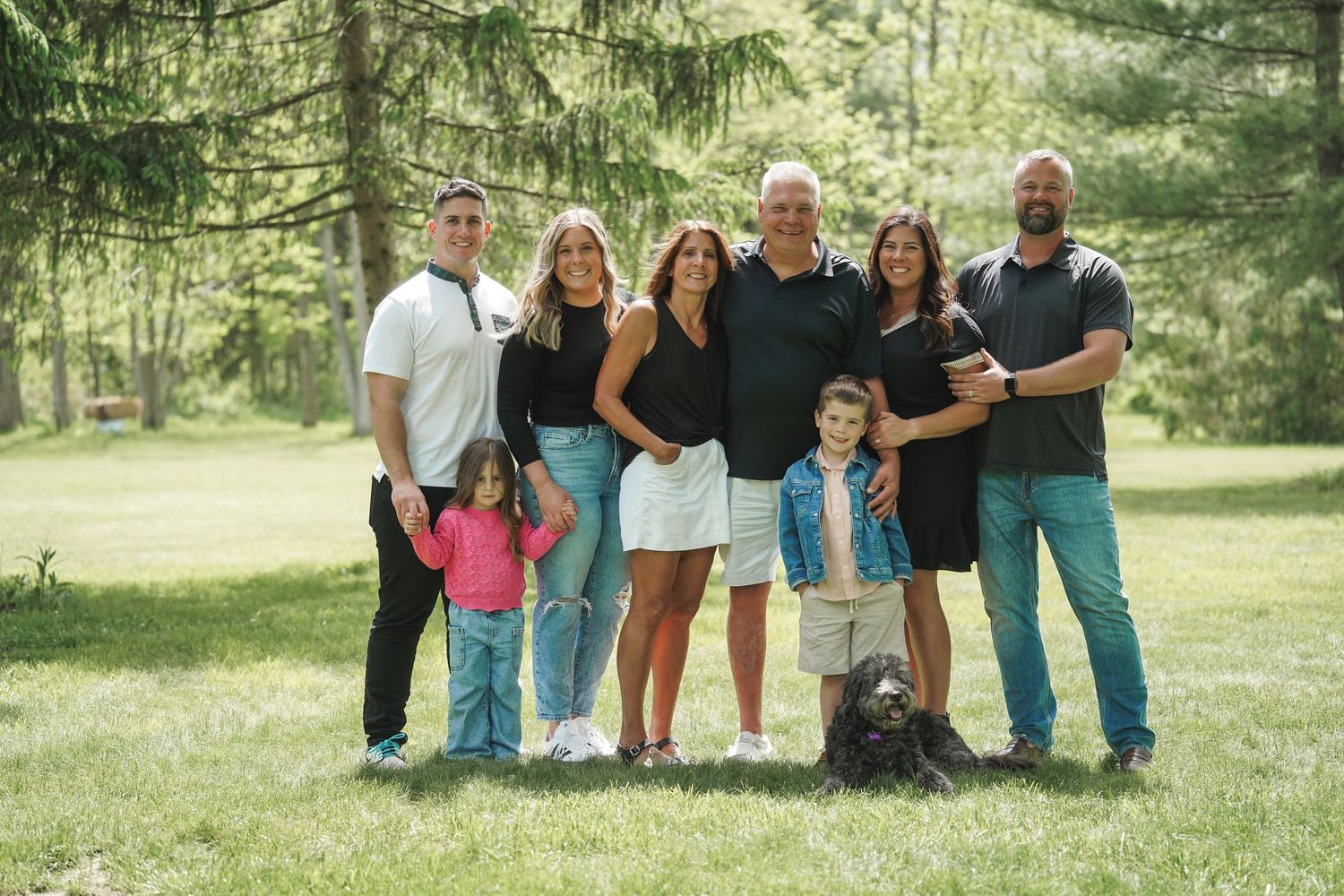 A multi-generational family group poses together outdoors on a grassy area with evergreen trees in the background.