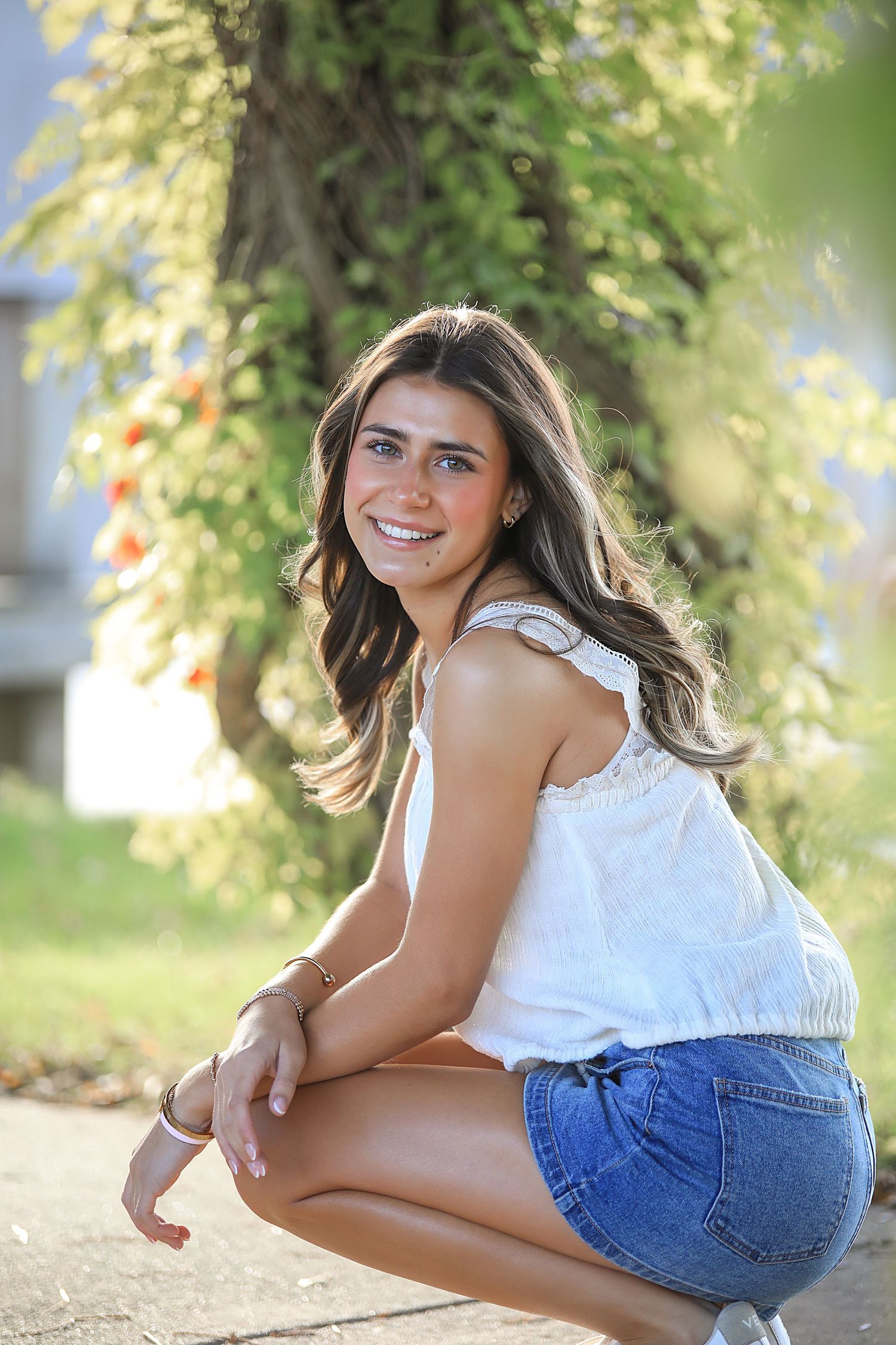 A person in a white sleeveless top and denim shorts poses outdoors on a sunny day with greenery in the background.