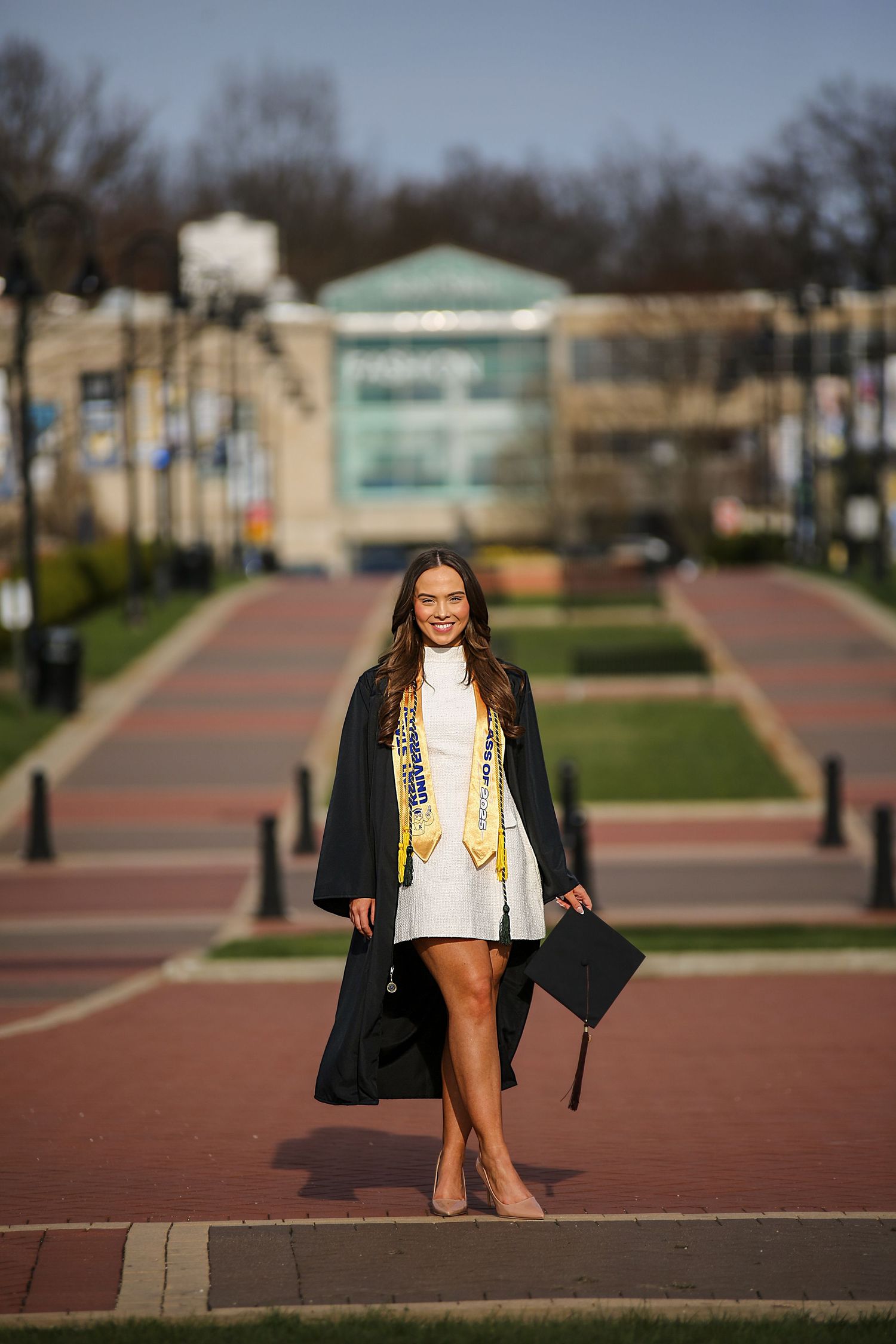 A graduate poses in academic regalia on a brick pathway during a sunny graduation photo session at a university campus.