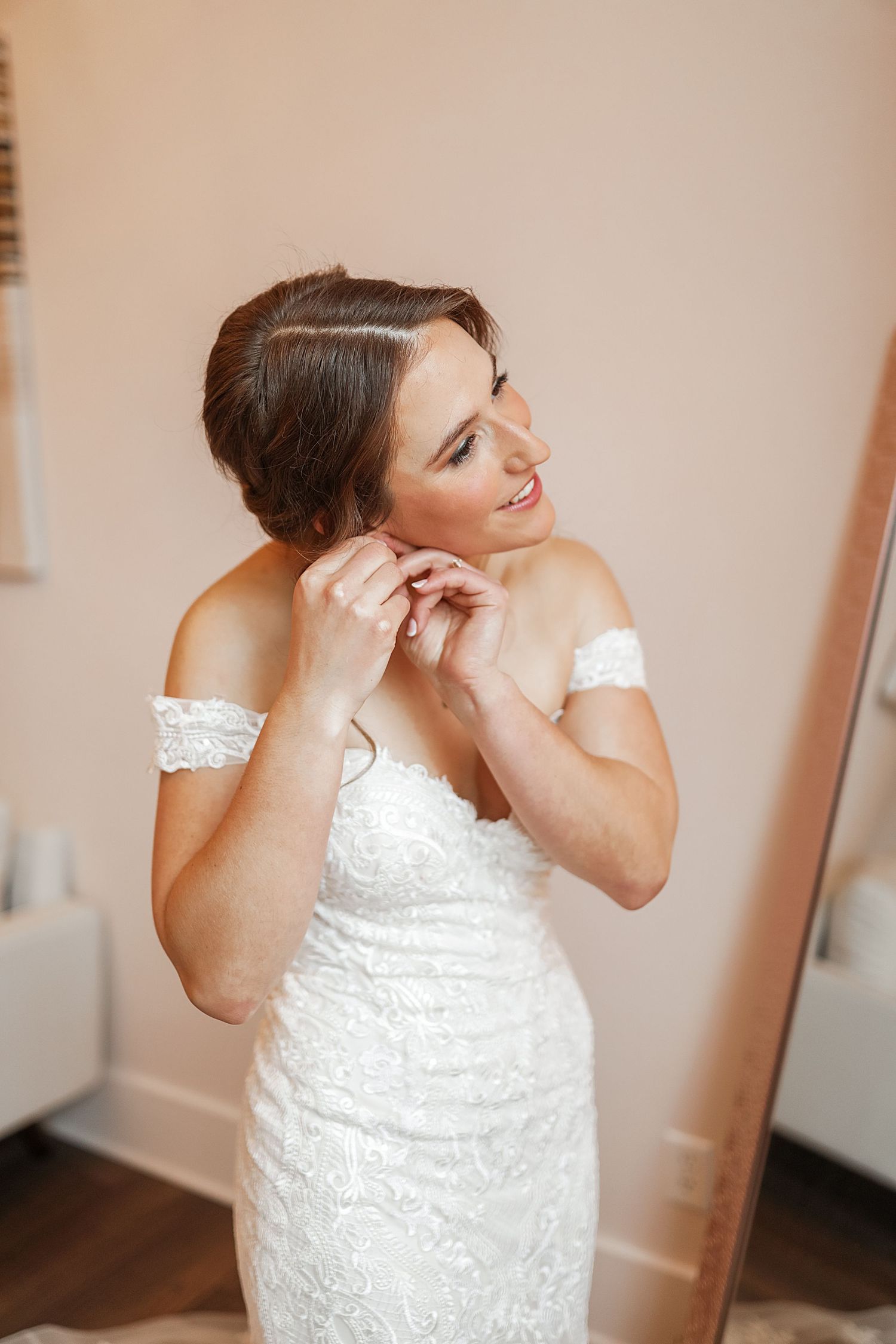 A bride in a white lace dress adjusts her hair while getting ready for her wedding day.