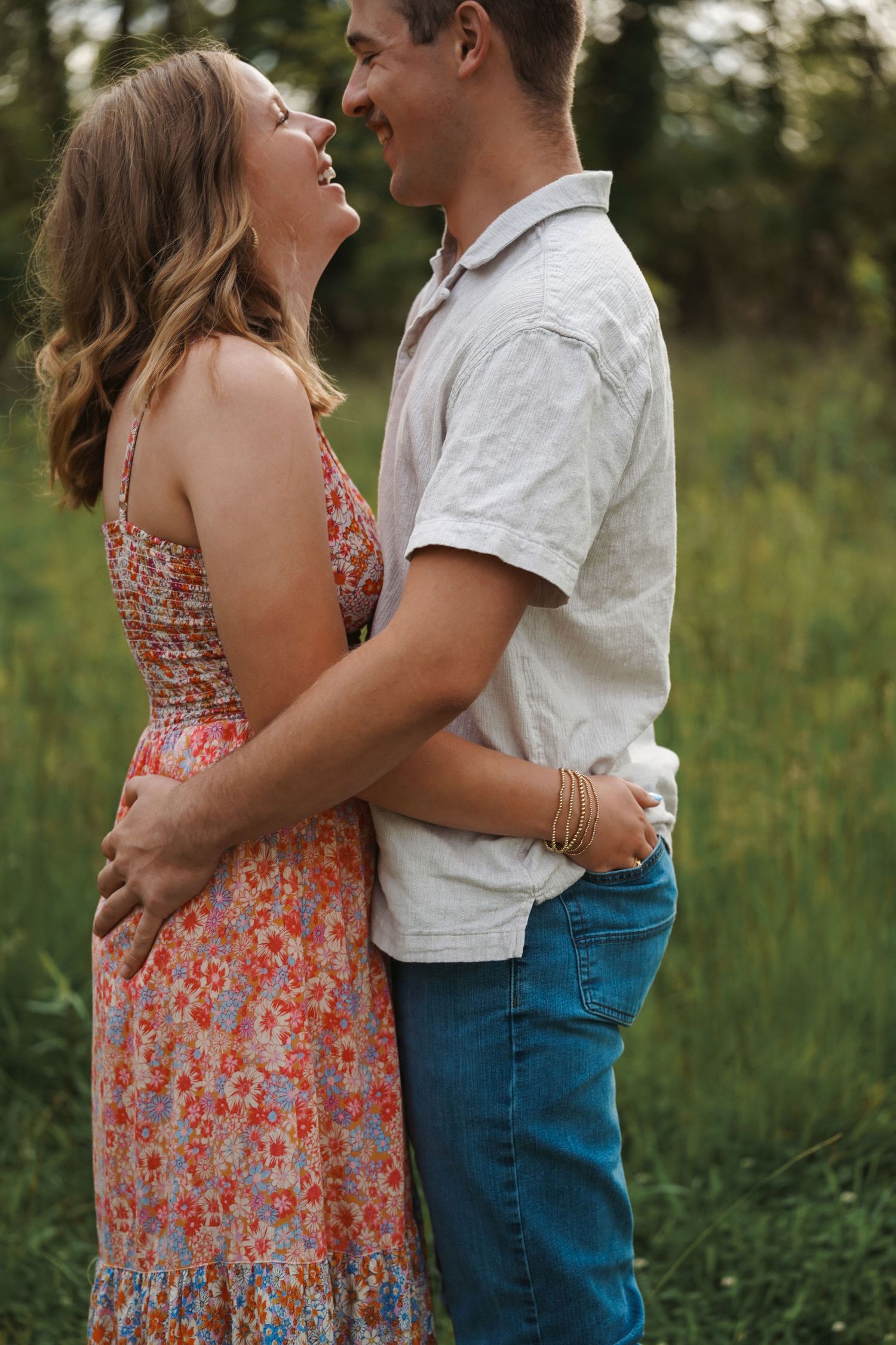 A couple in a floral dress and casual clothing embrace lovingly in a sunlit meadow during a romantic photoshoot.
