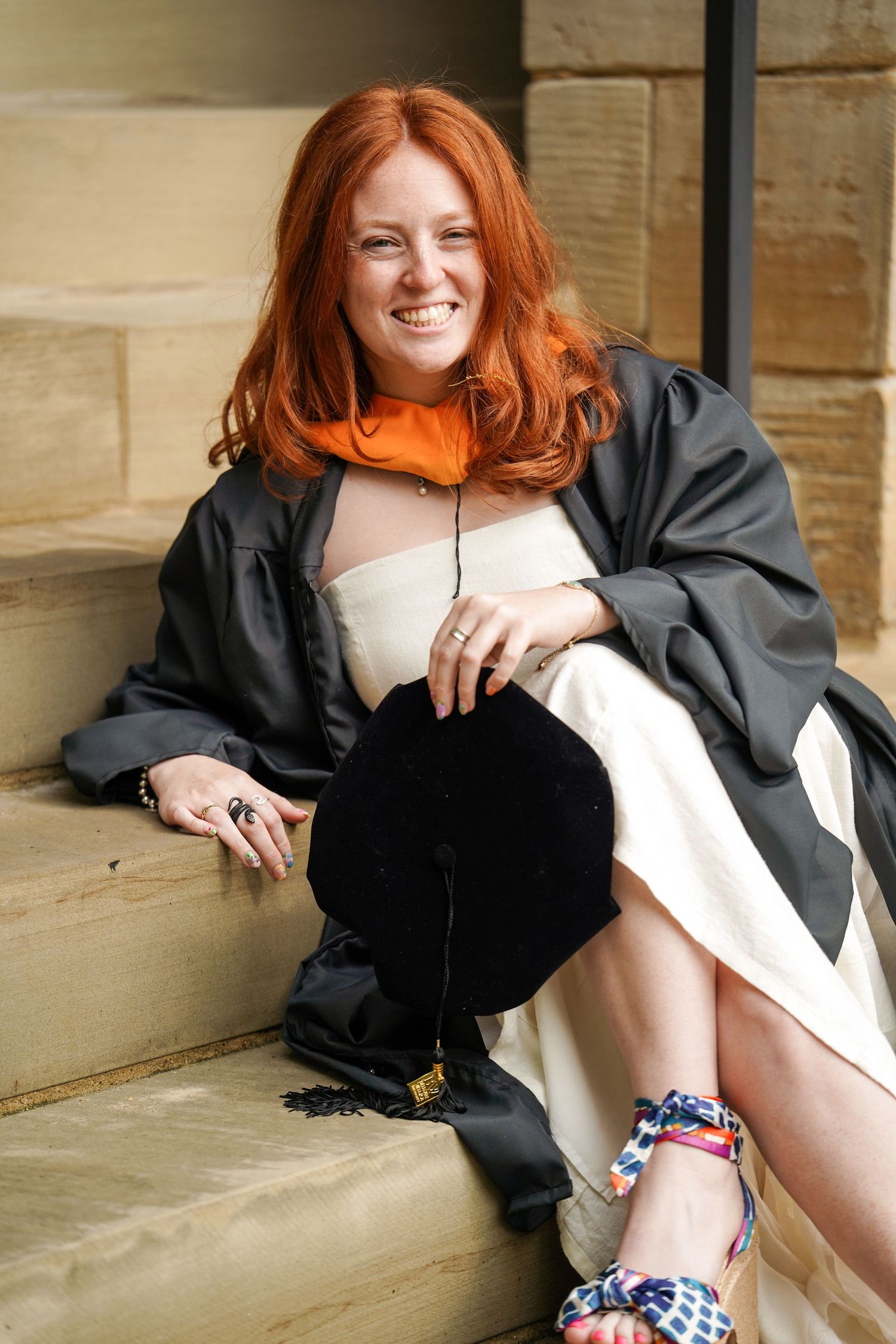 A graduate sits on stone steps wearing a black academic robe and colorful patterned shoes with bright red hair.