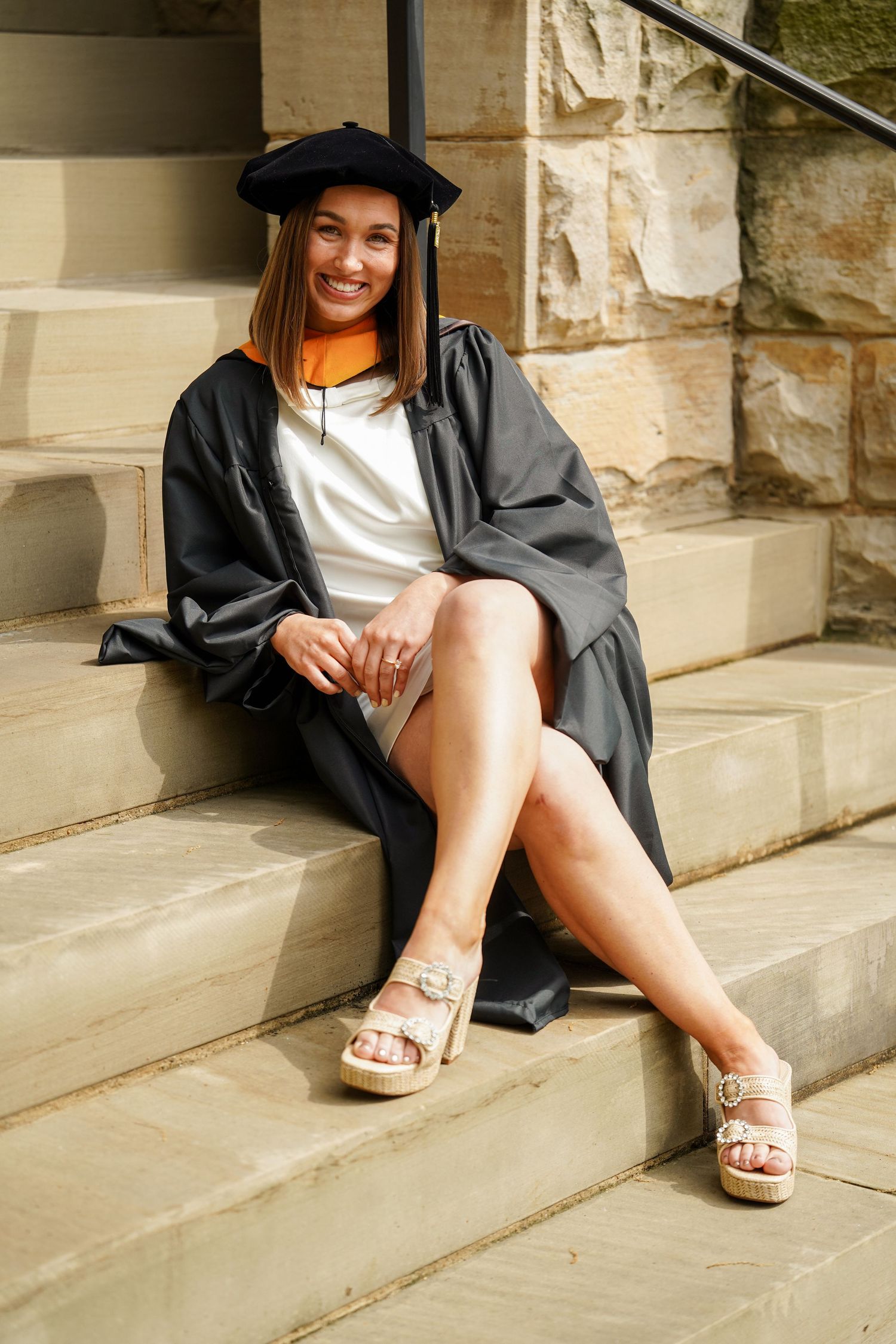 Graduate wearing traditional black cap and gown sits casually on stone steps while wearing platform sandals.