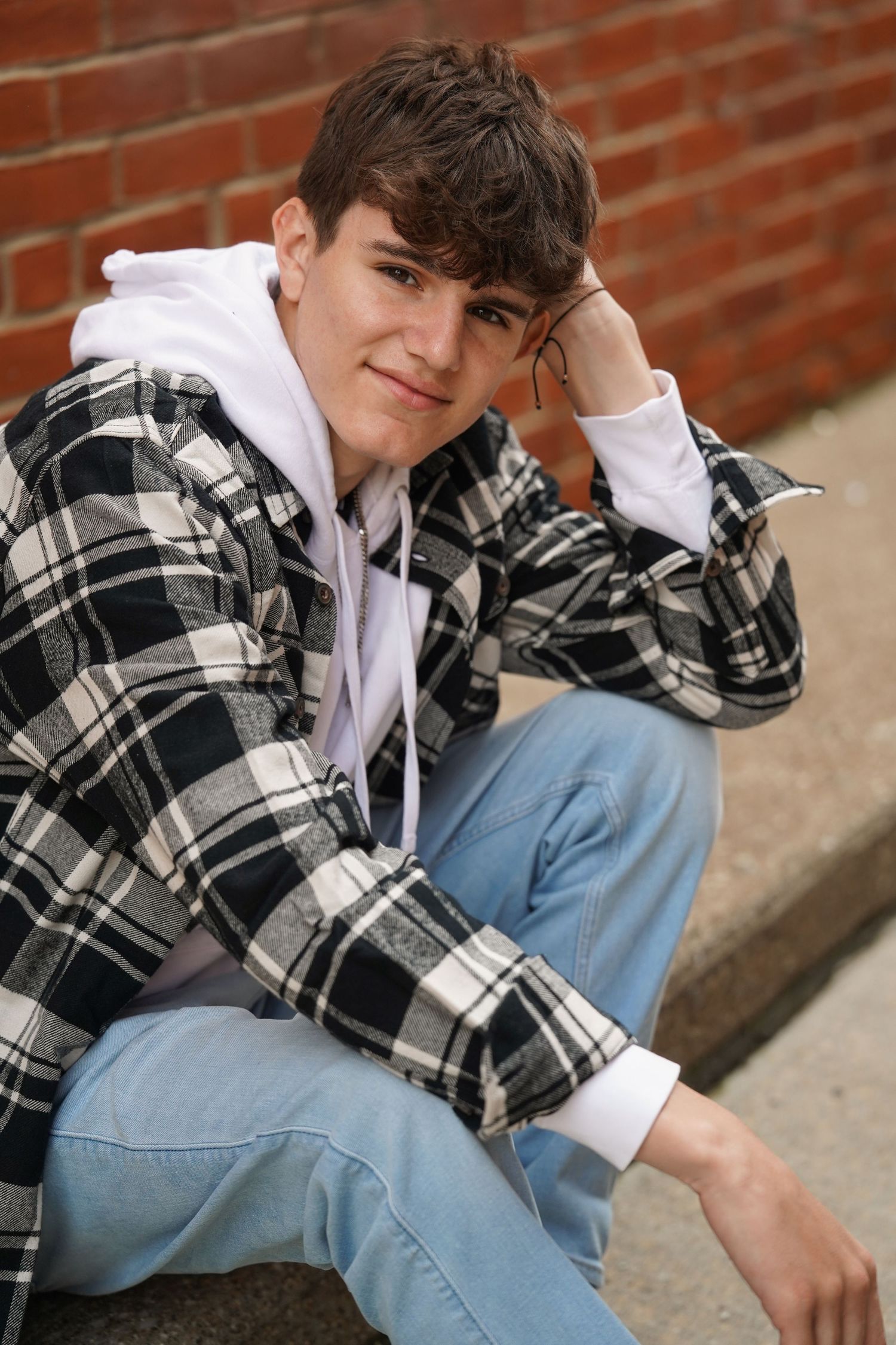 Young person in plaid flannel shirt and jeans poses casually against brick wall background.