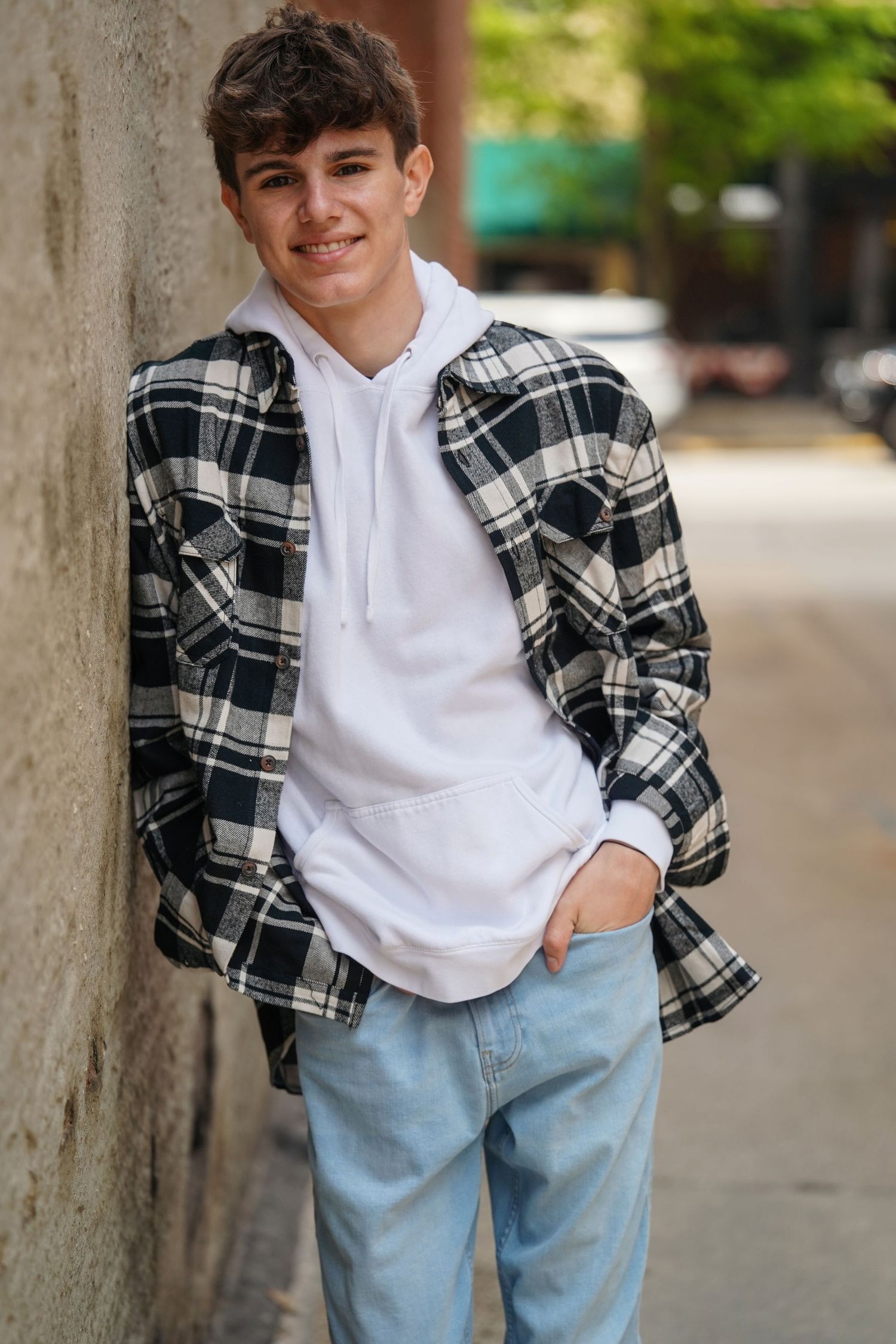 A person wearing a white hooded sweatshirt and plaid jacket poses casually against a concrete wall.