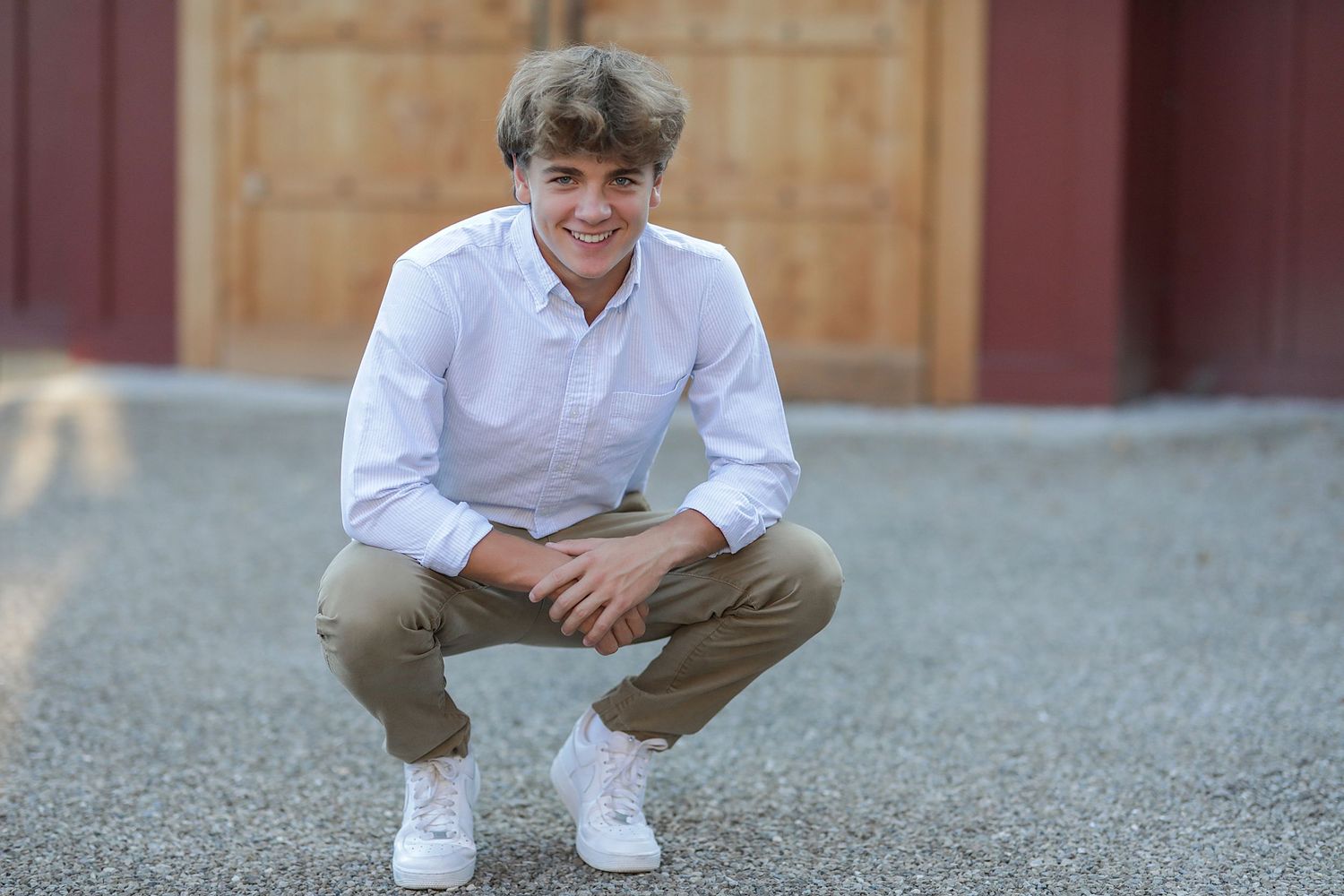 A young person in a white shirt and khaki pants sits casually on gravel near a red barn.