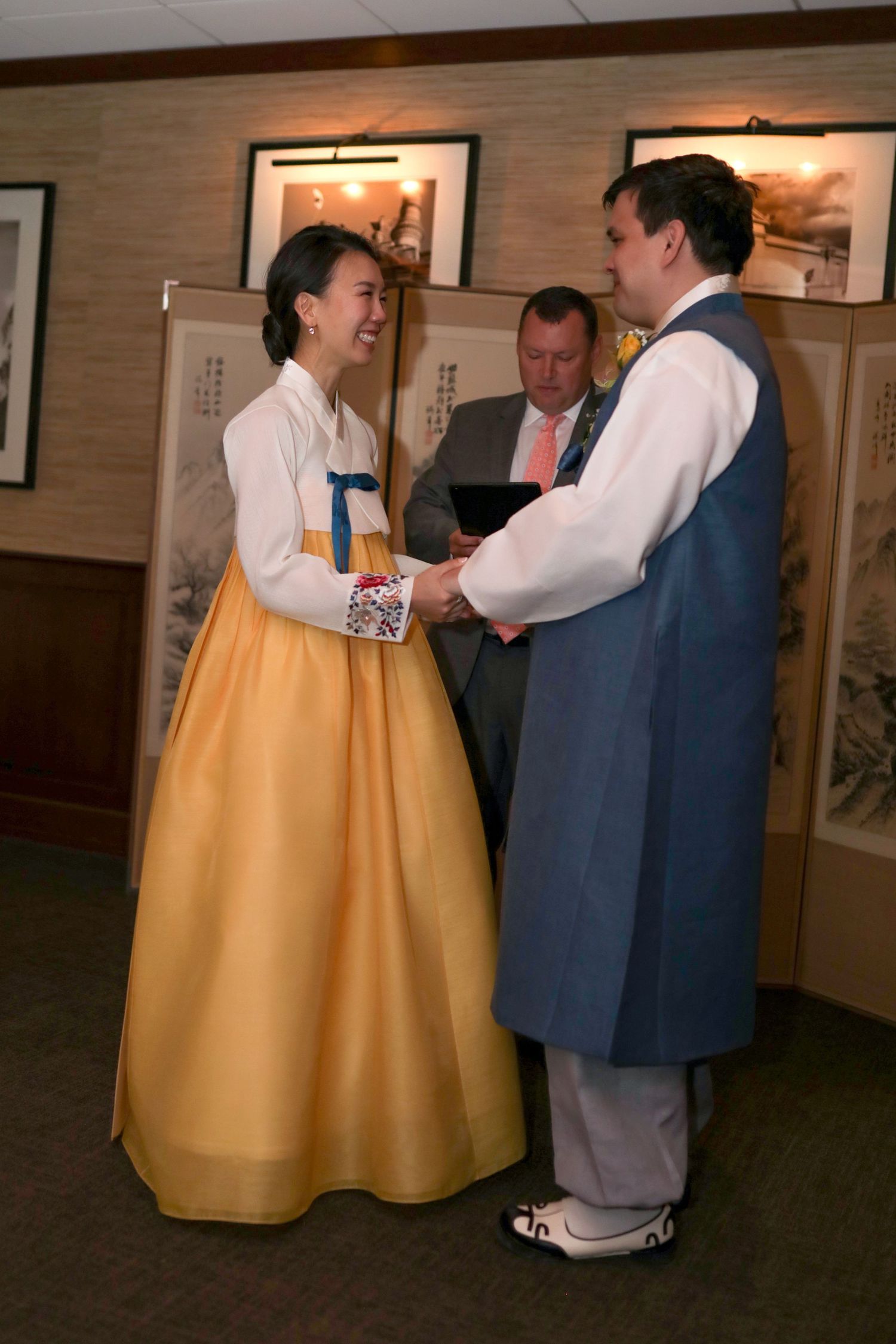 A couple wearing traditional Korean hanbok attire stand together while an officiant performs a ceremony in the background.