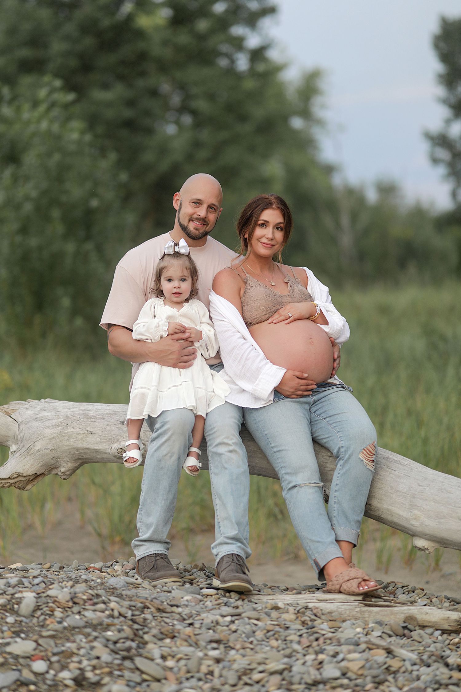 A family sits together on a driftwood log at a rocky beach while wearing coordinated casual outfits in neutral tones.