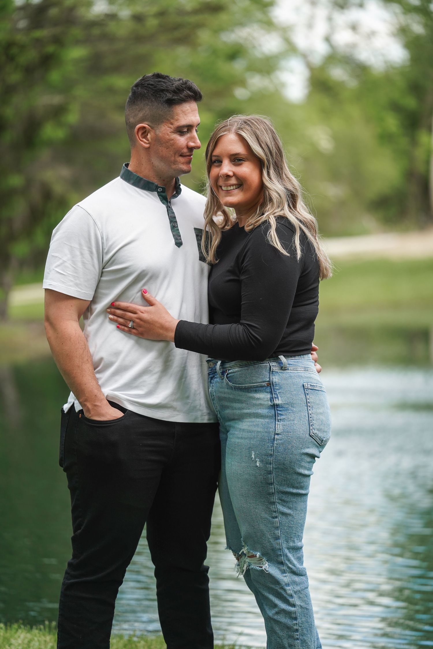A couple embraces by a pond on a sunny day, surrounded by lush greenery in a romantic outdoor setting.