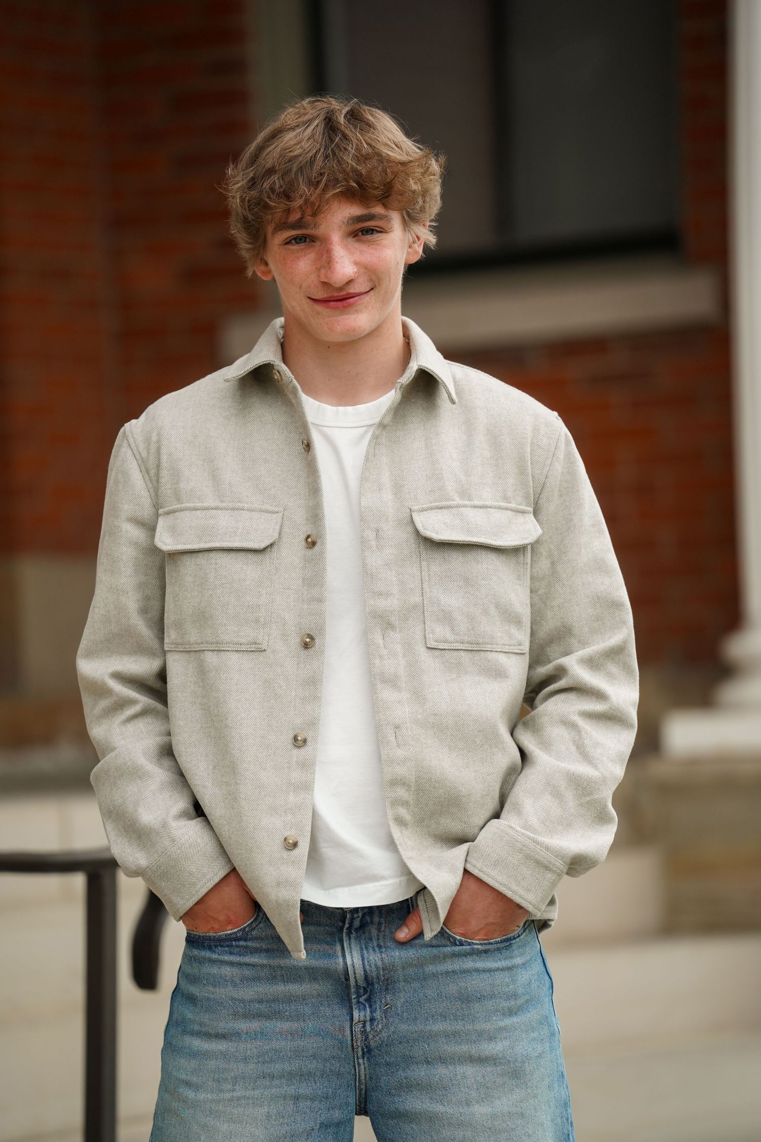 Person wearing light-colored button-up jacket and blue jeans poses against brick wall exterior in casual photo series.