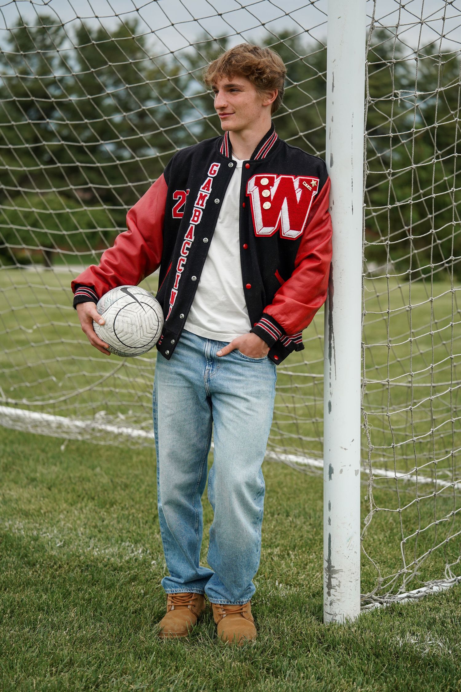 Sports player wearing a varsity jacket and jeans stands casually by a soccer goal net on a grassy field.