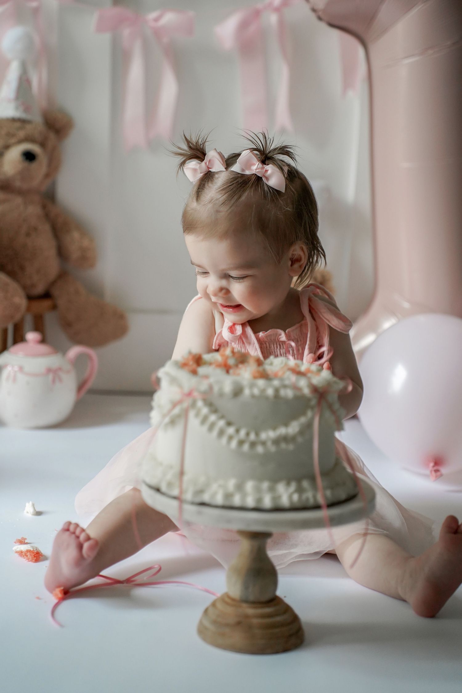 A baby in pink attire sits beside a delicate white birthday cake at a soft pink-themed celebration with balloons and teddy bear.