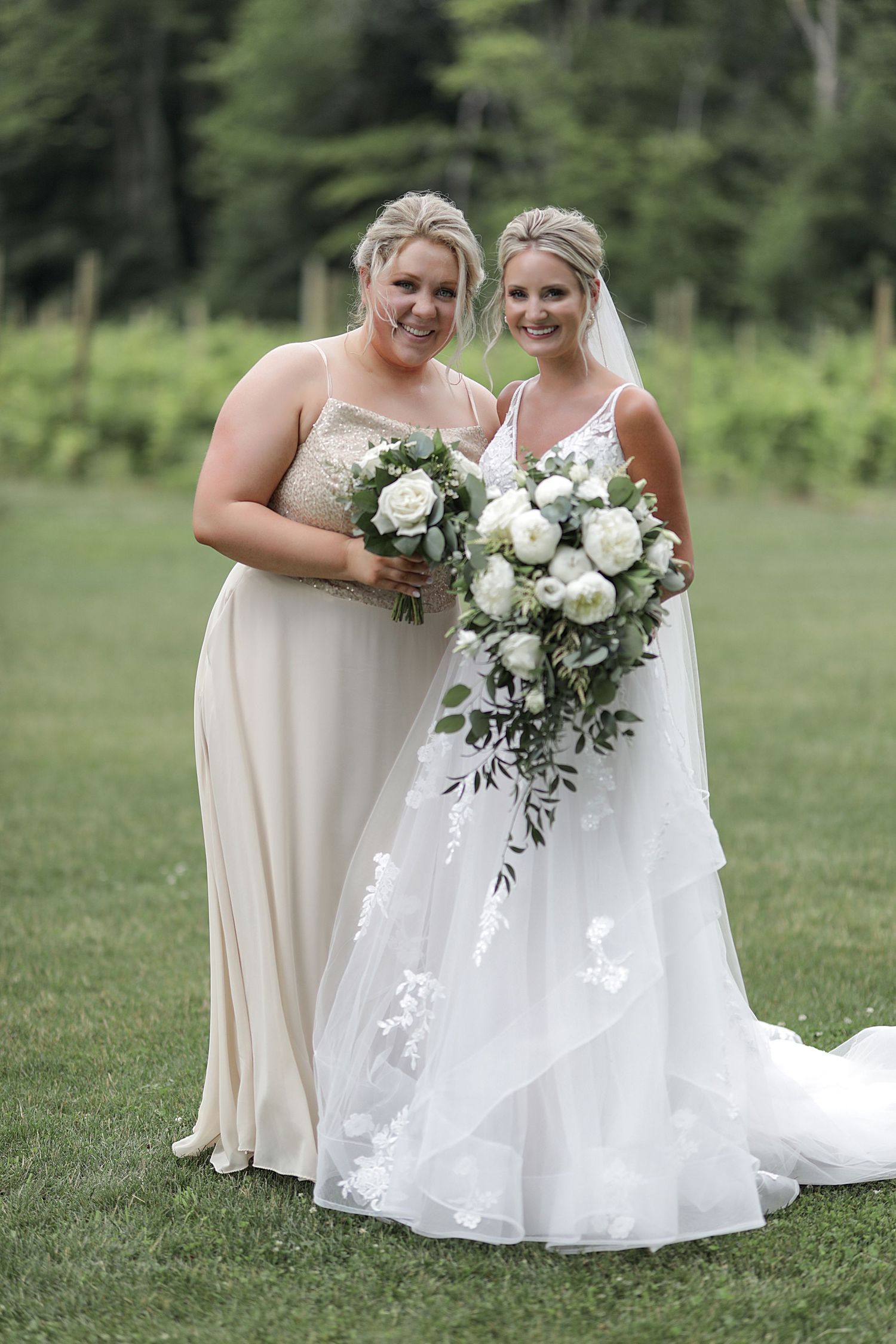 Bride and bridesmaids holding elegant white rose bouquets pose together on green lawn.