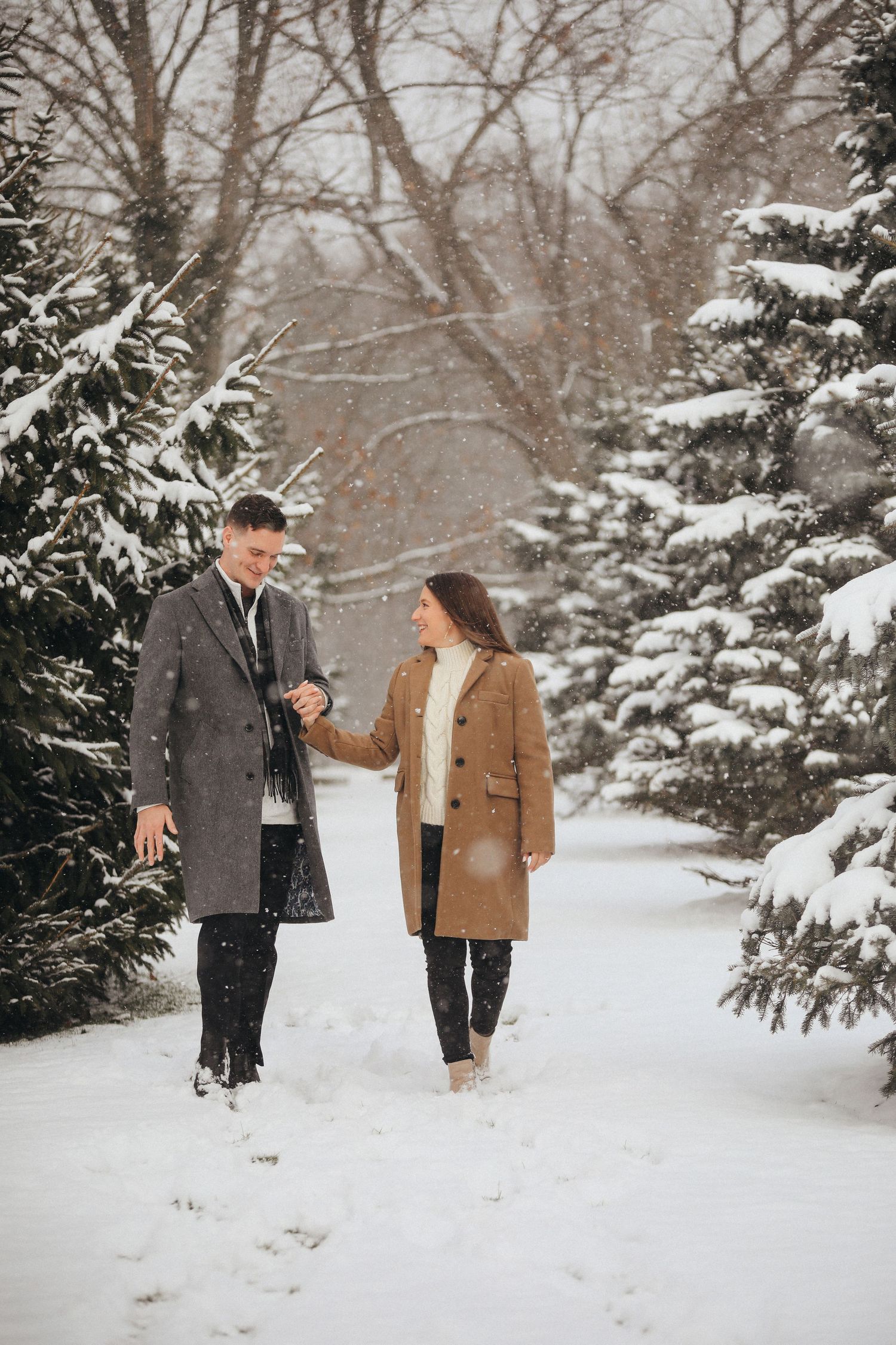 A couple walks together through a snowy winter forest path with evergreen trees covered in fresh white snow.