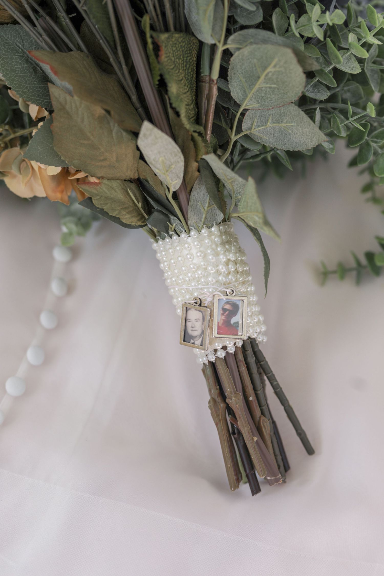 Wedding bouquet decorated with pearl charms holding loved ones' photos tied with white ribbons.