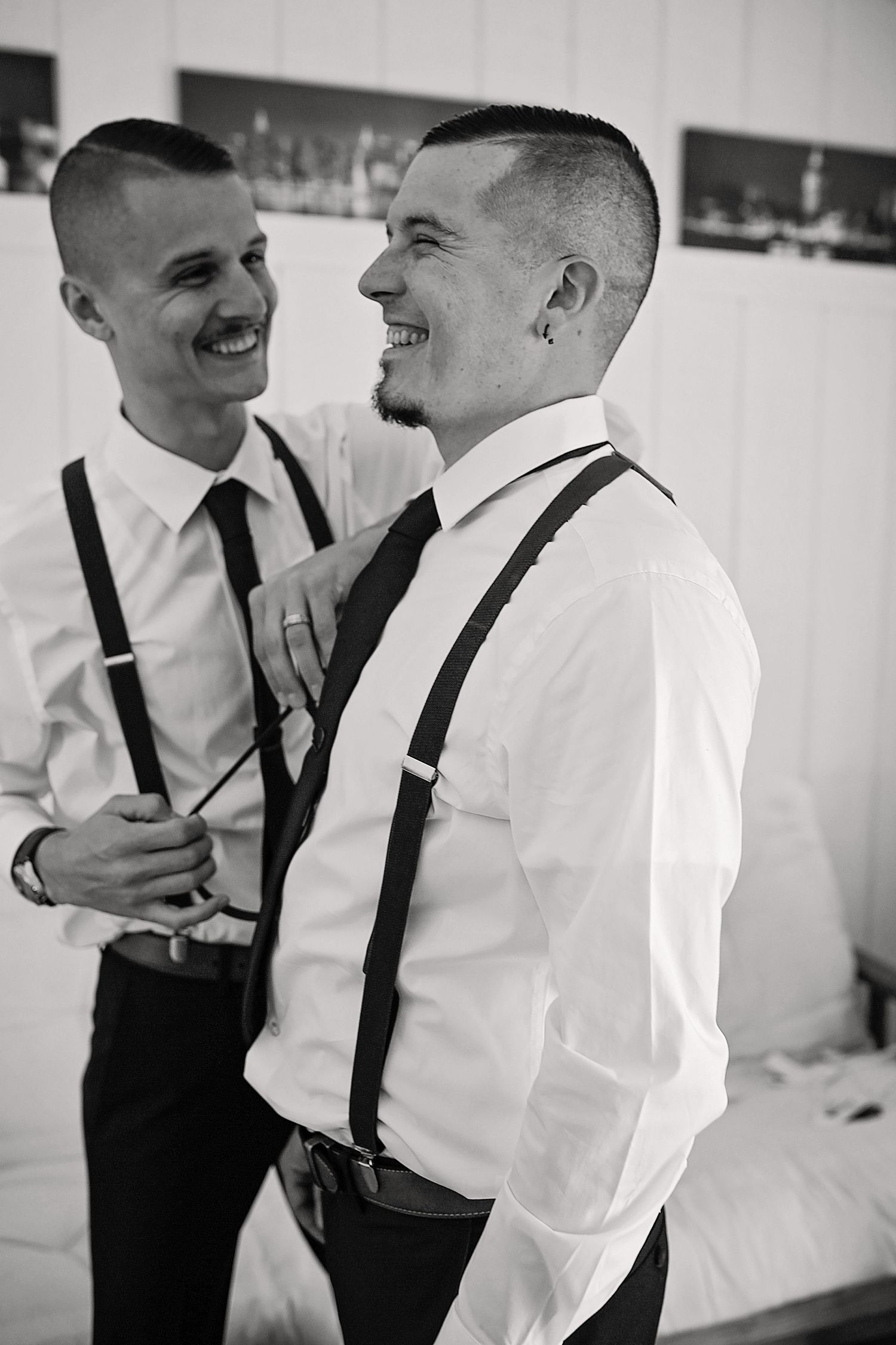 Two groomsmen in formal attire with suspenders share a candid black and white moment.