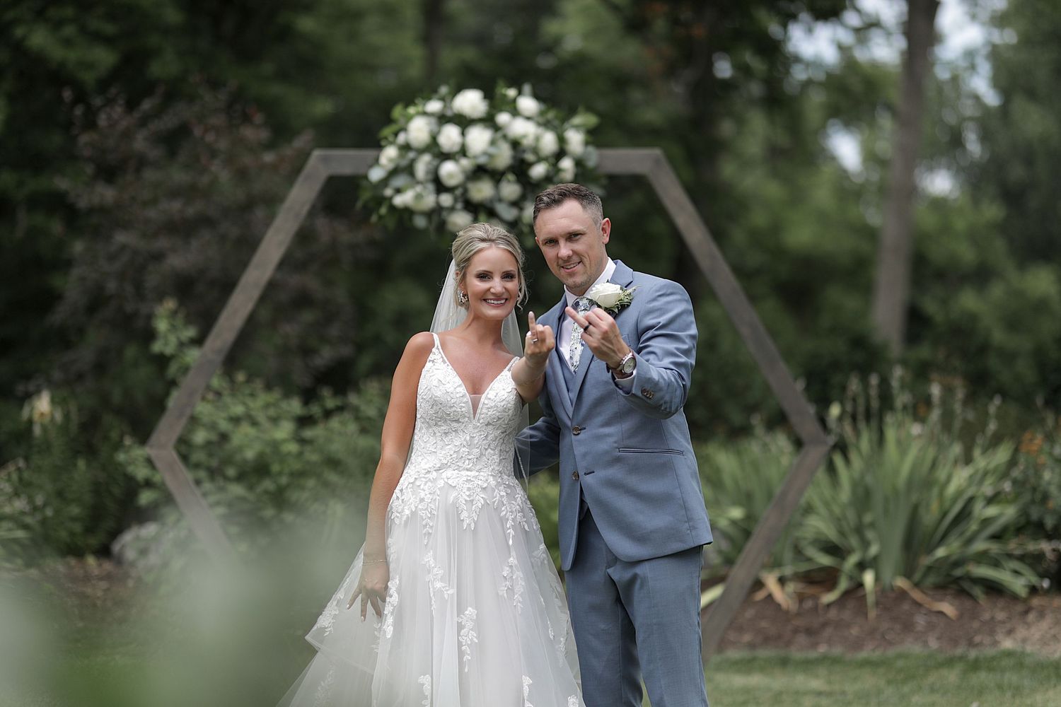 Wedding couple poses under wooden arch decorated with white florals in garden setting.