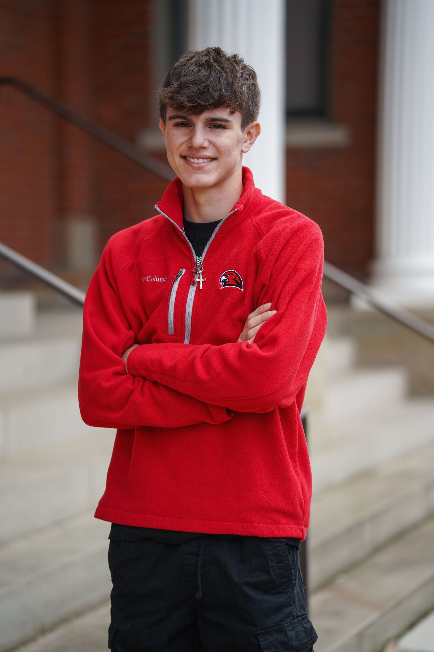 Student wearing a bright red fleece pullover and dark pants stands with arms crossed on stone steps.