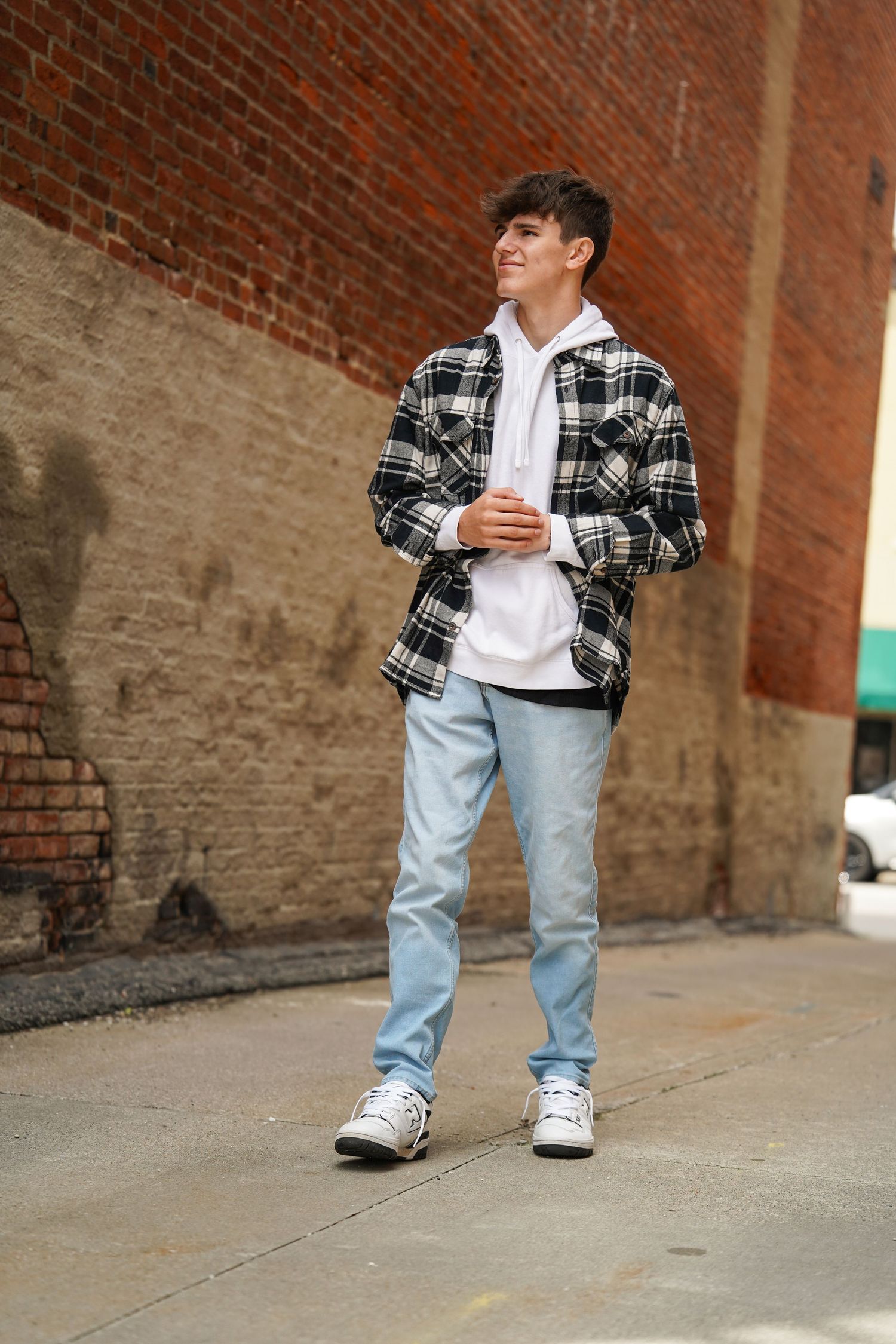 A person in a plaid flannel jacket and light jeans poses against a brick wall wearing white sneakers.