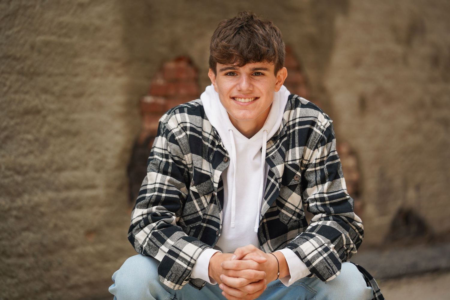 A person wearing a plaid flannel jacket and white hoodie sits and smiles at the camera against a tan wall.